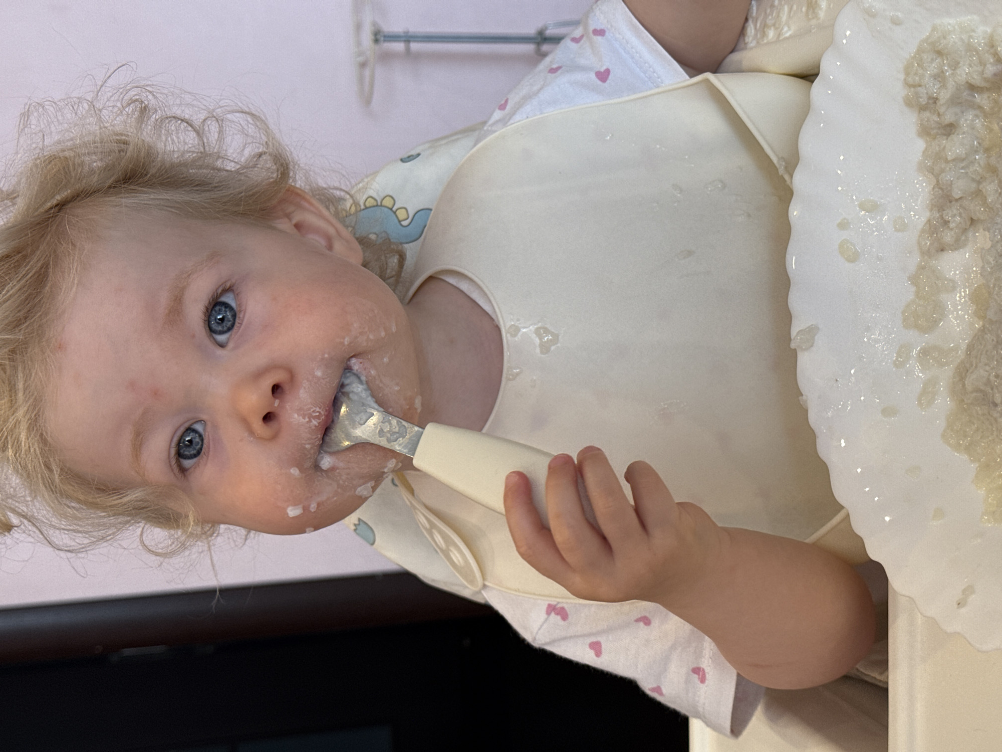 A young child with curly blonde hair and bright blue eyes is depicted in a close-up, rotated portrait, seemingly in the midst of a meal. The child looks directly at the viewer with an engaged expression, their face smeared with light-colored food, possibly oatmeal or porridge.
They are wearing a white long-sleeved shirt patterned with small pink hearts, and a pale silicone bib covers their front, also speckled with food. A faint blue dinosaur pattern is visible on an underlying garment. The child holds a spoon with a white handle, with food in their mouth and on the spoon.
To the child's right, a white plate or bowl is partially visible, filled with the same creamy, chunky food, which has also splattered onto the high chair tray and bib. The background is a plain, light-colored wall with a metallic fixture. The scene appears to be an indoor setting, likely a home in Vladimir, Russia, capturing a typical messy but endearing moment of a child learning to self-feed. There is no visible text in the image.