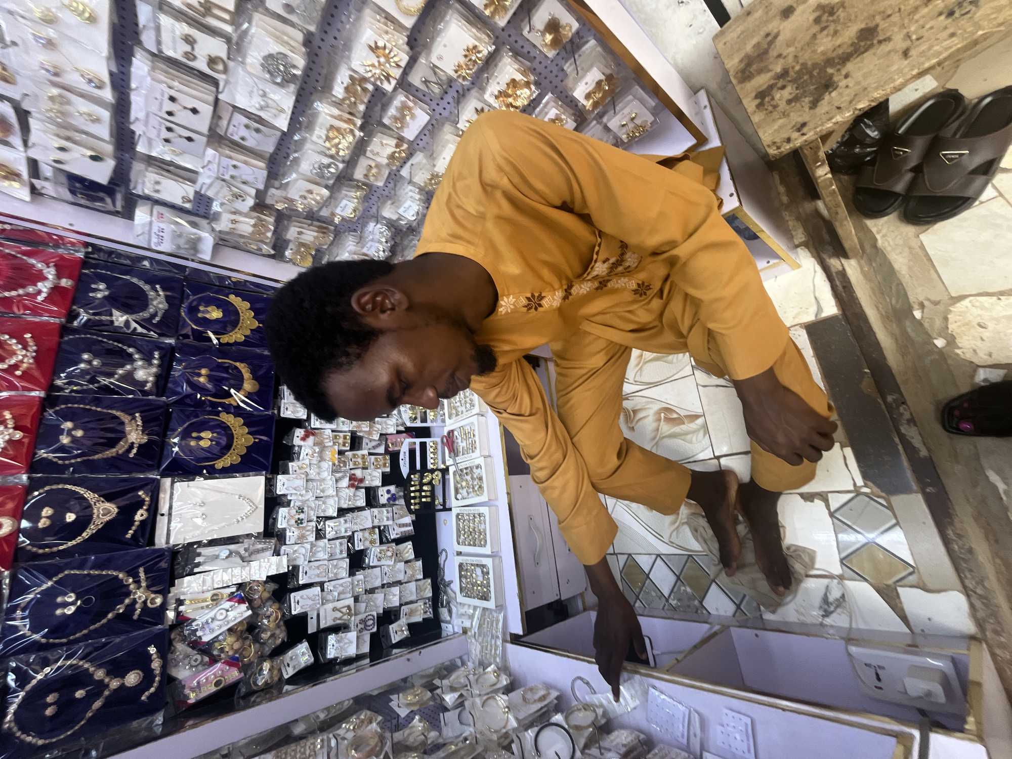 A man in a mustard-yellow traditional outfit is squatting in front of a stall filled with jewelry. The stall, located in Abuja, Nigeria, displays a wide variety of necklaces, earrings, bracelets, and rings. The jewelry is neatly arranged on shelves and in display boxes, some packaged in plastic. The man is looking down at the display and appears to be examining or organizing the items. The floor is tiled with a diamond pattern, and a wooden plank is visible nearby. The overall impression is of a small business or market stall.