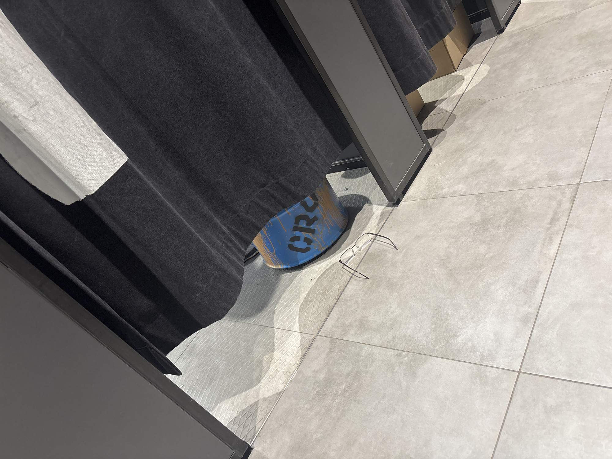 The image shows the floor of a retail space, possibly a fitting room, with light gray tiled flooring. A dark gray curtain hangs on the left side, partially obscuring a white textured material. In the center of the frame, a blue cylindrical object with "CRS" printed in black is visible. A pair of glasses rests on the floor near the blue object. A cardboard box is partially visible behind the curtain. The environment appears to be indoors. The location is identified as Astana, Kazakhstan.