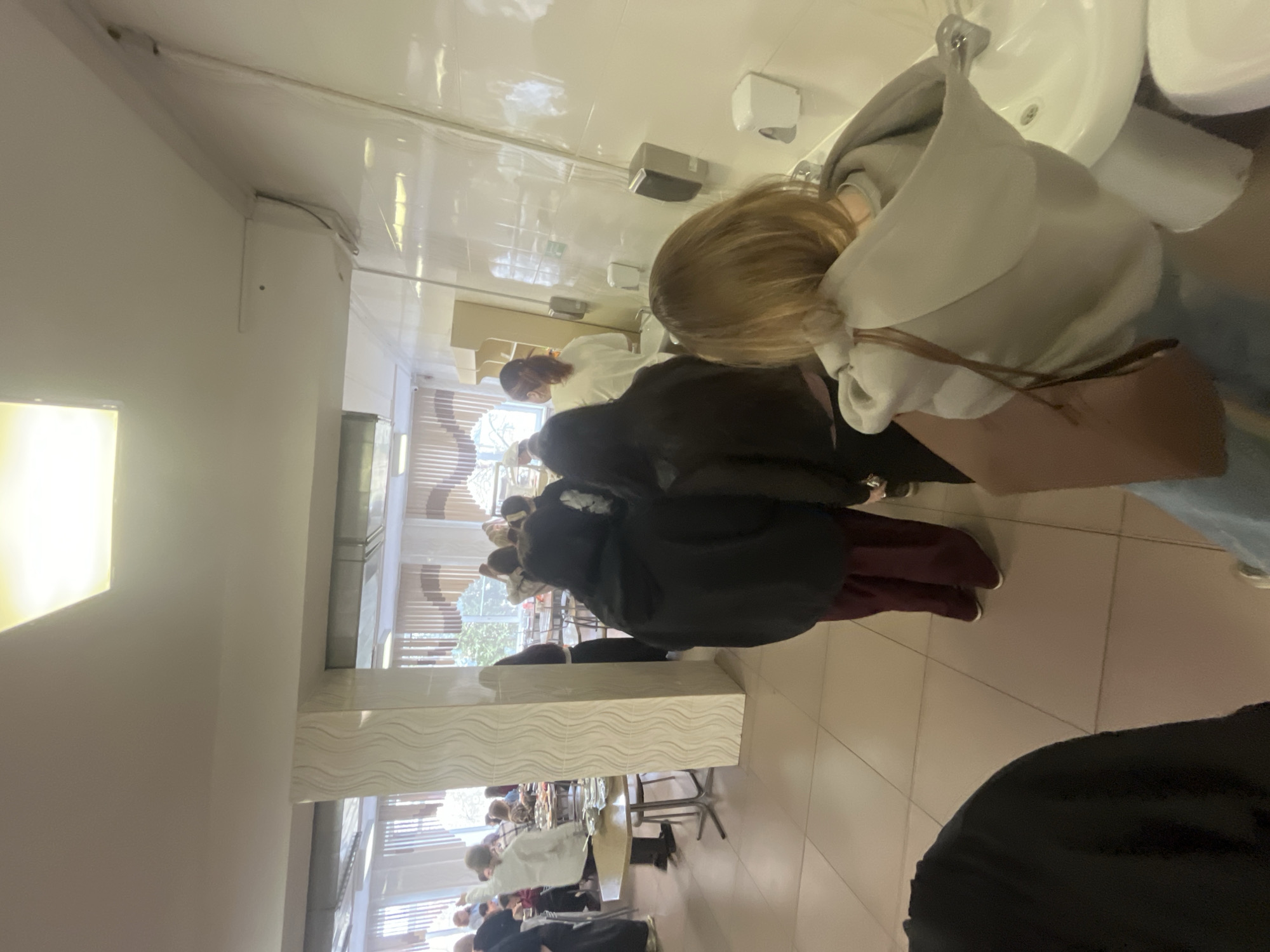 Here is a forensic description of the image:
**General Description:** The image appears to be taken indoors, likely in a public or semi-public space. There are multiple people present, primarily women, engaged in different activities. The image is slightly skewed, possibly taken from a phone.
**People:**
* A woman with blonde hair is washing her hands at a sink. She wears a gray hoodie, blue jeans, and a brown crossbody bag.
* Several individuals are standing in line, some wearing dark coats or jackets.
* Further in the background, multiple people are seated at tables, likely eating or socializing.
**Objects:**
* Several white sinks and associated fixtures are visible.
* Hand soap dispensers are mounted on the wall near the sinks.
* Tables and chairs are present in the background.
* A large, decorative column with textured surface is located on the left.
**Actions/Interactions:**
* People are washing hands.
* People are standing in line, possibly waiting to wash hands or get food.
* People in the background are seated and engaged in conversation, potentially eating.
**Environment:**
* The space appears to be a canteen.
* The floor is tiled in a light color.
* Large windows are in the background.
* The location could be Saint Petersburg based on the general architectural style.
**Geographic Location:**
Given the architectural characteristics visible through the window and the interior design, the location could potentially be within Saint Petersburg, Russia, however, further information is necessary for confirmation.