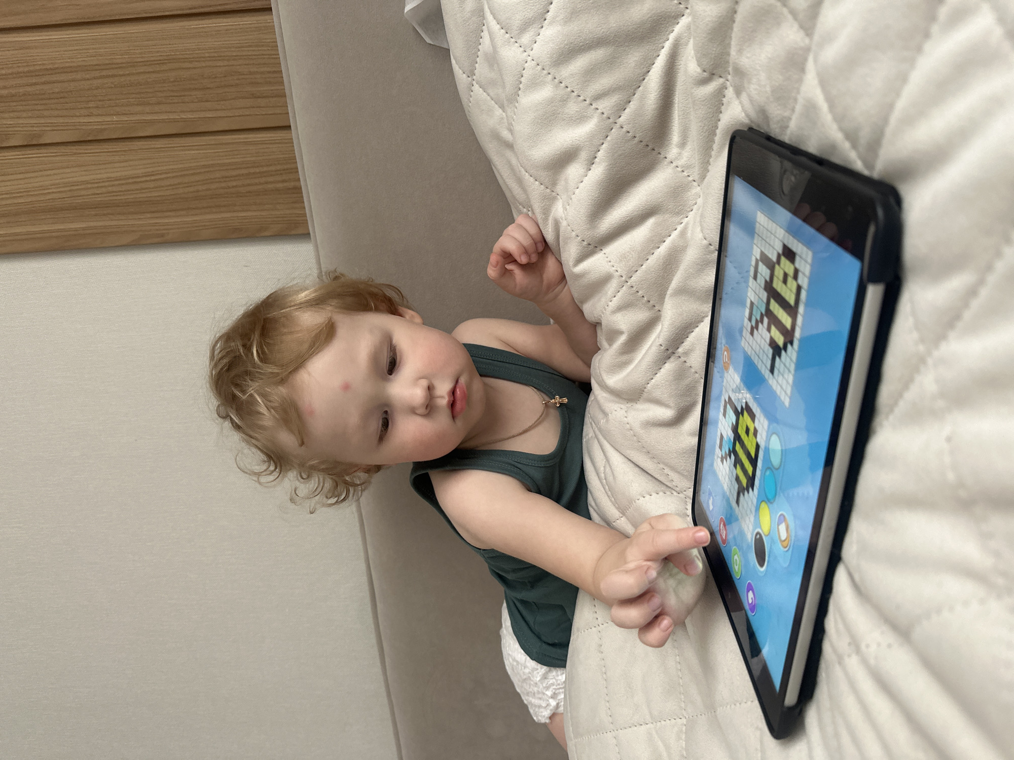 A toddler with curly blonde hair, wearing a green tank top and a diaper, is sitting on a white quilted surface, possibly a bed. The toddler is looking at a tablet screen, which displays a colorful game or app, and is touching the screen with their right index finger. The location is unknown.