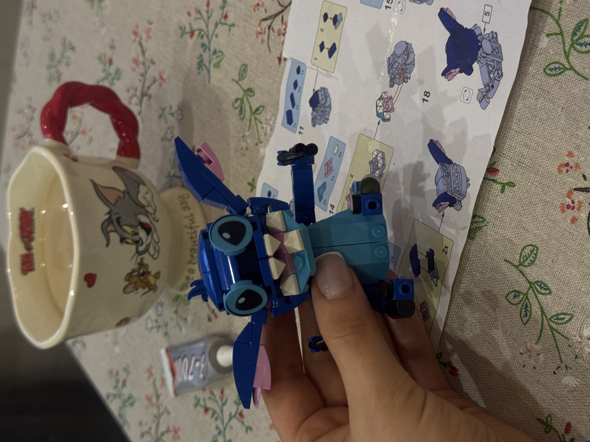 A close-up shot captures a hand holding a nearly-completed LEGO Stitch figure over a surface with scattered items. The LEGO figure, predominantly blue, features large, round eyes, white teeth, and pink accents on its ears. A LEGO instruction manual lies adjacent, displaying diagrams of the figure's construction stages. To the left, a Tom and Jerry mug with a red handle adds a touch of whimsy to the scene. A small tube of glue, marked "3-70K," is visible nearby. The background consists of a tablecloth or placemat with a floral pattern. The location is not Moscow, Russia.