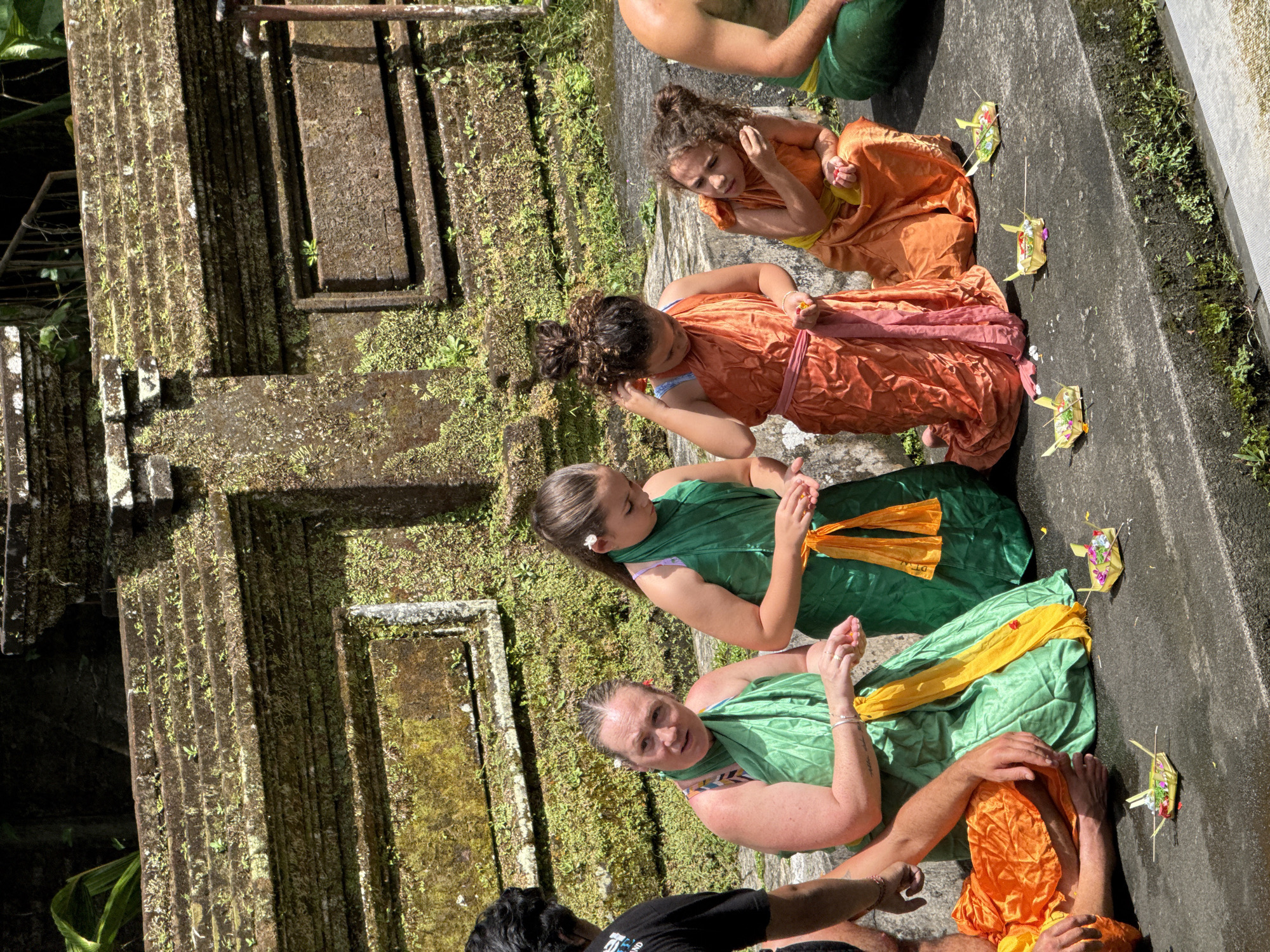 The image captures a group of individuals participating in a cultural or religious ceremony outdoors, characteristic of Tegallalang, Indonesia. Four people are seated or kneeling on a gray concrete surface, with their hands often clasped or in a posture of prayer, facing toward moss-covered stone structures.
In the central and lower portions of the frame, two adults wear bright green traditional attire, likely sarongs, with vibrant yellow and orange sashes. One woman in green has a white flower tucked behind her ear and looks towards the camera with a gentle expression. Behind them, two young girls and another adult are dressed in flowing orange or reddish-orange garments, some featuring pink sashes. The girls have curly hair, often tied up.
Arranged on the ground beside the participants are several small, square-shaped offerings, known as Canang Sari, made from woven palm leaves and containing flowers and other small elements, which are common in Balinese Hindu rituals. A person in a black t-shirt is partially visible in the bottom foreground, and another individual in an orange garment is seen lower right.
The background consists of imposing, multi-layered stone structures, heavily overgrown with green moss and small plants, suggesting an ancient temple or traditional water architecture. A dark channel or pathway runs along the base of these structures. The scene is illuminated by bright natural sunlight, indicating midday or early afternoon conditions with clear skies. The overall mood appears focused and reverent. No decipherable text is visible on any objects or clothing.