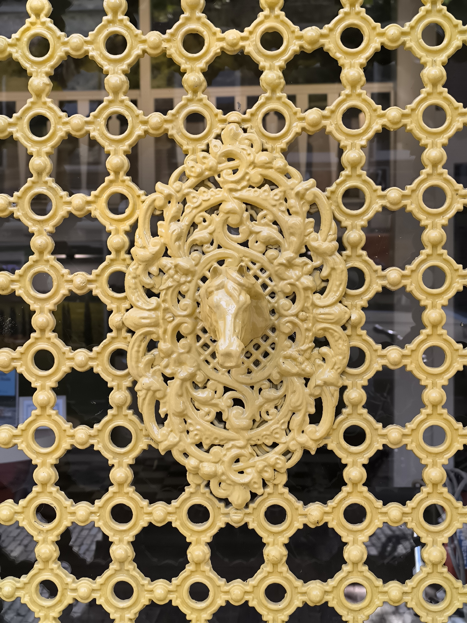 A close-up, eye-level, vertical shot captures a decorative, light yellow metal grate, likely part of a window or fence, in Alkmaar, Netherlands. The grate is designed with a repeating geometric pattern of interconnected circles and dots. Centered on the grate is an ornate, oval-shaped embellishment featuring a relief of a horse's head with flowing mane, framed by intricate scrollwork and floral motifs. The metal appears to be cast and has a smooth, slightly glossy finish. Through the openings of the grate, reflections of a dark background and some indistinct building structures are visible, suggesting the grate is mounted on a window or facade. The overall impression is one of historical or architectural ornamentation.