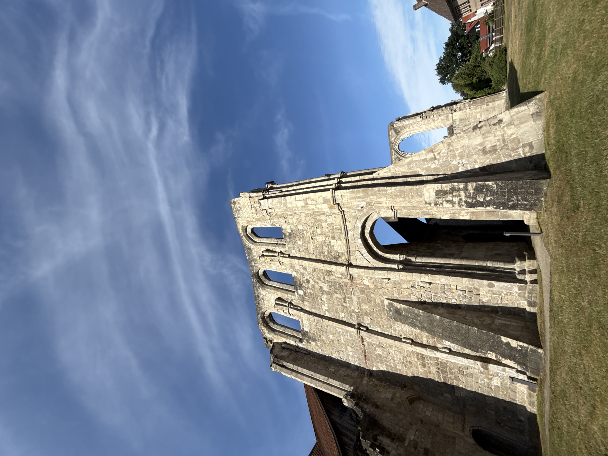 In Walkenried, Germany, an outdoor shot captures the imposing Gothic ruins of a former monastery against a clear blue sky dotted with wispy white clouds. The main subject is the weathered stone facade of the structure, characterized by its tall, arched windows, some of which are gaping openings to the sky. Buttresses support the massive stone walls, casting long shadows on the grass-covered ground below. To the left, a section of a red-tiled roof is visible, indicating that part of the monastery complex may still be intact or has been restored. In the background, a few houses with red roofs and green trees form a pastoral setting. The scene is serene and evokes a sense of history and architectural grandeur.