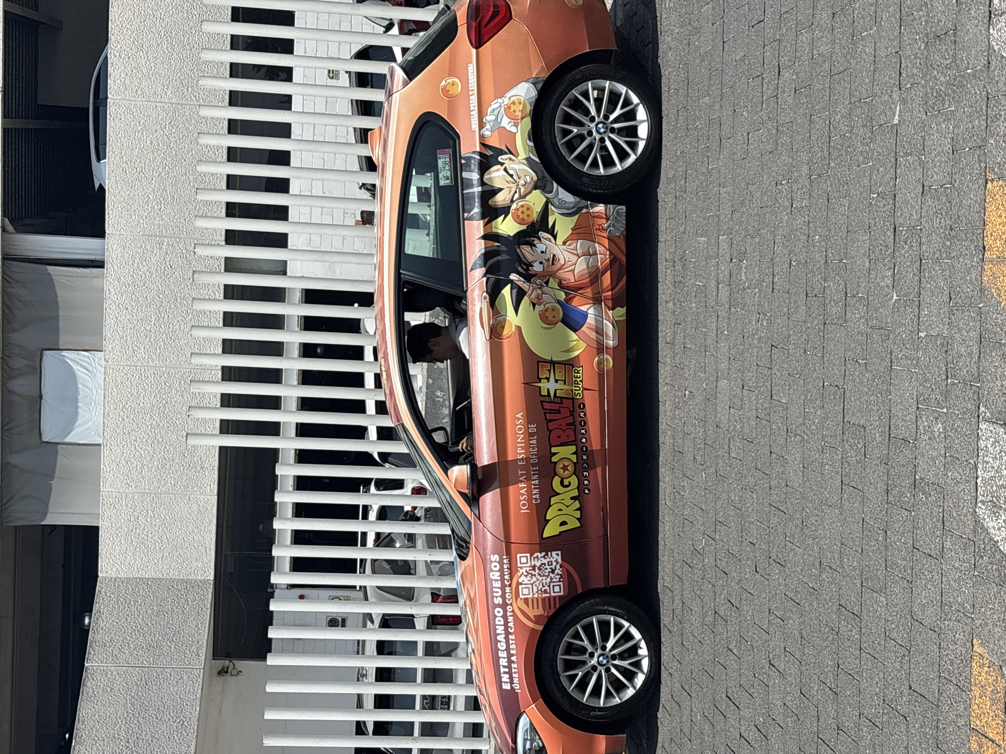 The image depicts an orange car with a "Dragon Ball Super" theme parked on a gray-tiled surface, likely a street or parking area. The car features images of Goku and Vegeta, along with Dragon Ball Super branding. Text on the car includes "JOSAFAT ESPINOSA, CANTANTE OFICIAL DE DRAGON BALL SUPER" and "ENTREGANDO SUEÑOS, ¡ÚNETE A ESTE CANTO CON CAUSA!" A QR code is also visible.
The car is parked next to a building with a white fence. Several other vehicles are visible through the gaps in the fence. A person is inside the car.
The car's license plate appears to read "WF-258-8". The location is indicated on the license plate to be Huixquilucan de Degollado, Mexico.