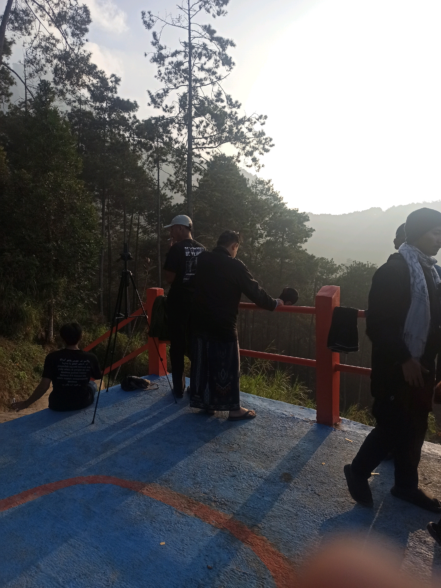 The image depicts a group of people at an outdoor overlook, likely a tourist spot, with a view of rolling hills and dense pine forests, characteristic of the higher altitudes surrounding Cileunyi, Indonesia. The sky is bright and hazy, suggesting it might be early morning or late afternoon. In the foreground, a blue-painted concrete platform is visible, marked with red lines.
On the platform, there are several individuals. To the left, a person is seated with their back to the camera, wearing a black t-shirt with text. A tripod with a camera is positioned nearby, indicating potential photography or videography activities.
In the center of the frame, two men are standing. One, wearing a white cap and a black t-shirt, appears to be smoking and looking towards the view. The other man, also in dark clothing, is facing away from the camera, with a dark sarong-like garment around his waist. A red railing runs along the edge of the platform, with a few items, possibly clothing, draped over it.
To the right of the frame, another individual is standing, wearing a black jacket and a light-colored scarf. Their face is partially visible, and they are looking to the side. The background is dominated by lush green trees, primarily pine trees, receding into a misty haze that obscures the distant landscape. The lighting suggests a natural outdoor environment, possibly a viewpoint or resting area along a scenic route in the Cileunyi area.