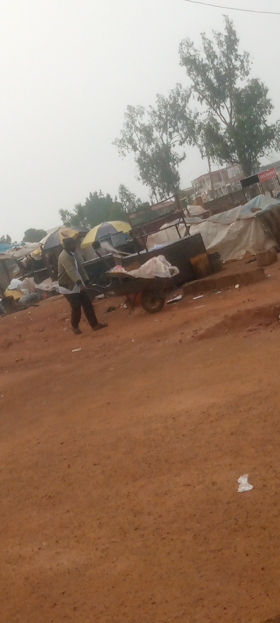 The image shows an outdoor scene in Gura Top, Nigeria, depicting a market environment. In the foreground, a person dressed in a light-colored shirt and dark pants is pushing a two-wheeled cart laden with various items, including white plastic bags and what appear to be bundled goods. The cart has a dark-colored basin and a visible wheel on the right side. The ground is covered in reddish-brown dirt.
In the middle ground, several market stalls are visible. These stalls are covered with tarpaulins and umbrellas, some of which are yellow and blue. Objects are stored under these coverings, and some appear to be bags of goods. A sign that reads "POS SERVICE" is visible on a red background in the background, suggesting the presence of a service point.
The background features several trees with sparse foliage, some of which appear to be eucalyptus trees, and buildings. The sky is overcast and hazy, contributing to a muted lighting condition. The overall impression is of a bustling, informal market setting.