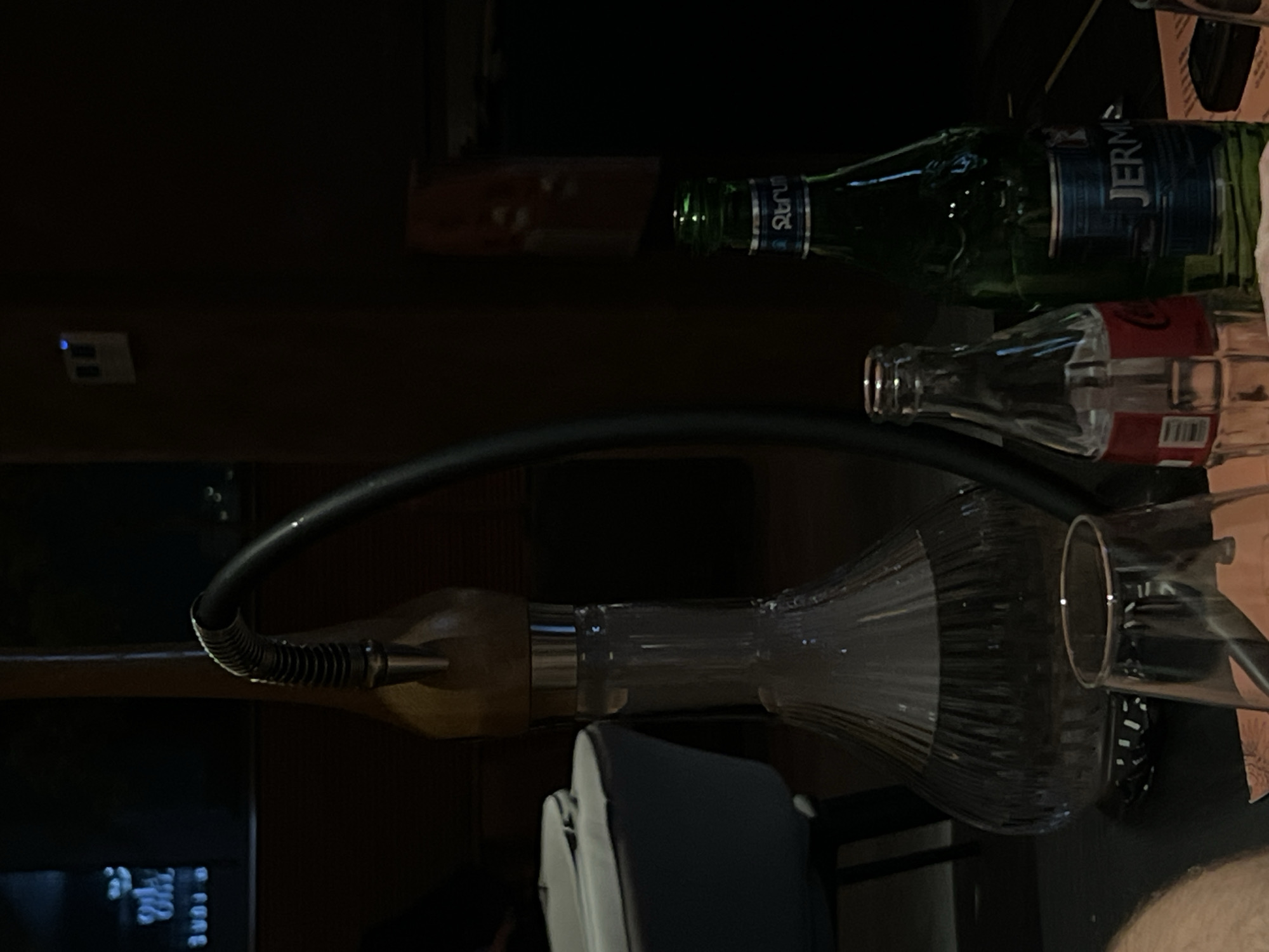 The image shows a dimly lit interior setting, likely a lounge or bar, possibly in Yerevan, Armenia, considering the presence of a "Jermuk" mineral water bottle. A hookah, with its distinctive glass base, hose, and wooden mouthpiece, is prominently displayed in the foreground. An empty Coca-Cola glass bottle lies next to the hookah. Above, a bottle of "Jermuk" mineral water is positioned horizontally, indicating it may have been used recently. A section of a dark wooden panel is faintly seen above the bottle of "Jermuk". A light switch can be seen to the left. The partial view of a seat suggests a person was or is present.