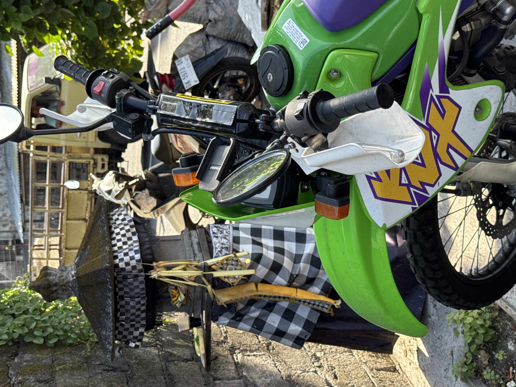 The image shows a parked green and purple Kawasaki dirt bike in an urban environment, possibly in Bali, Indonesia given the black and white checkered cloth and offerings on the bike. The bike has handguards, a small digital display, and a lockable fuel cap. Other vehicles are nearby. The background includes foliage, part of a building, and what appears to be a stone structure or monument near the left side of the frame. The photograph is taken at ground level.