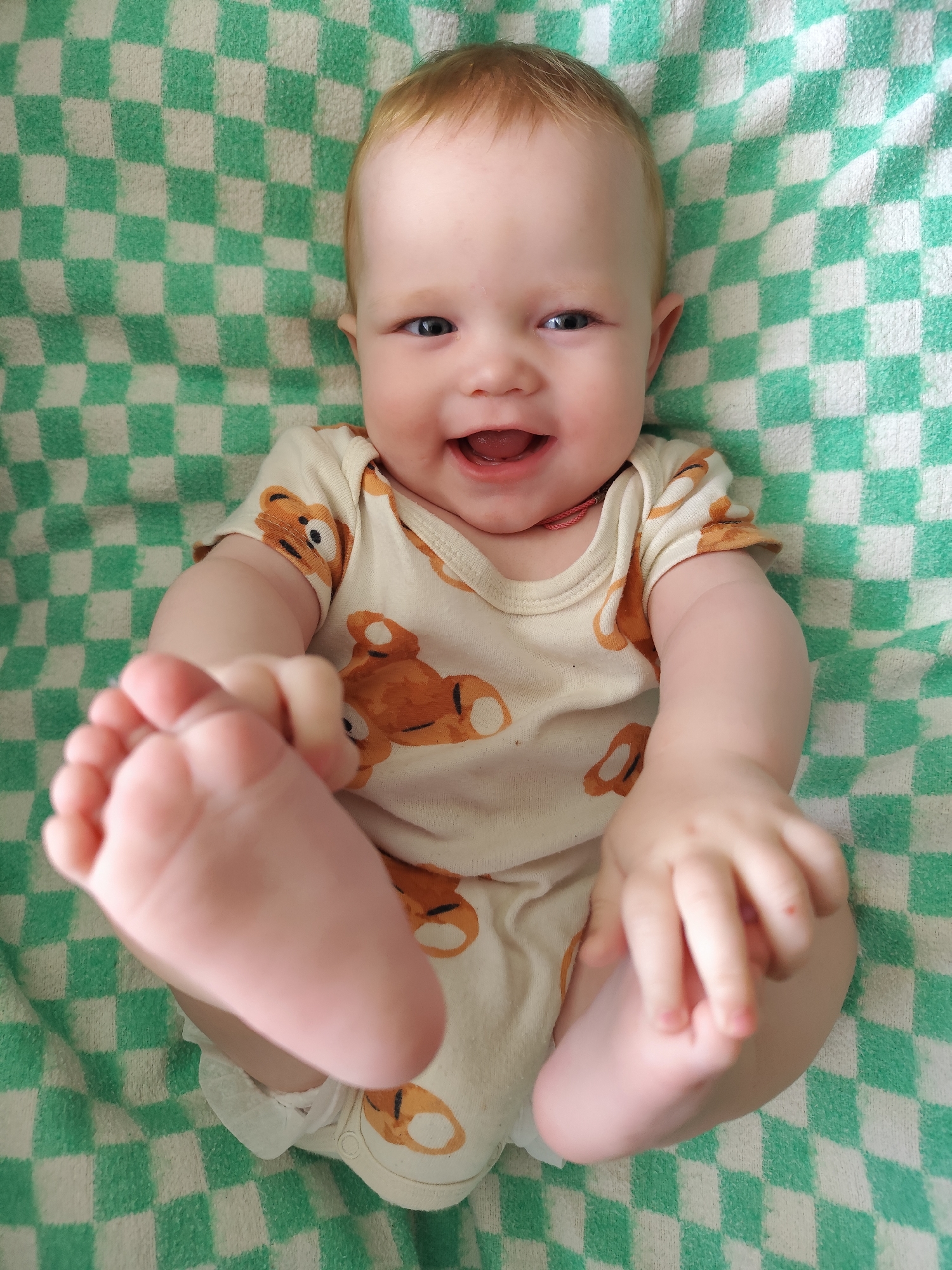 A baby is lying on a green and white checkered blanket. The baby is wearing a light-colored onesie with brown teddy bear patterns. The baby is smiling and has a foot raised in the air and is holding it with one hand. There is no indication of Фоки, Russia in this image.