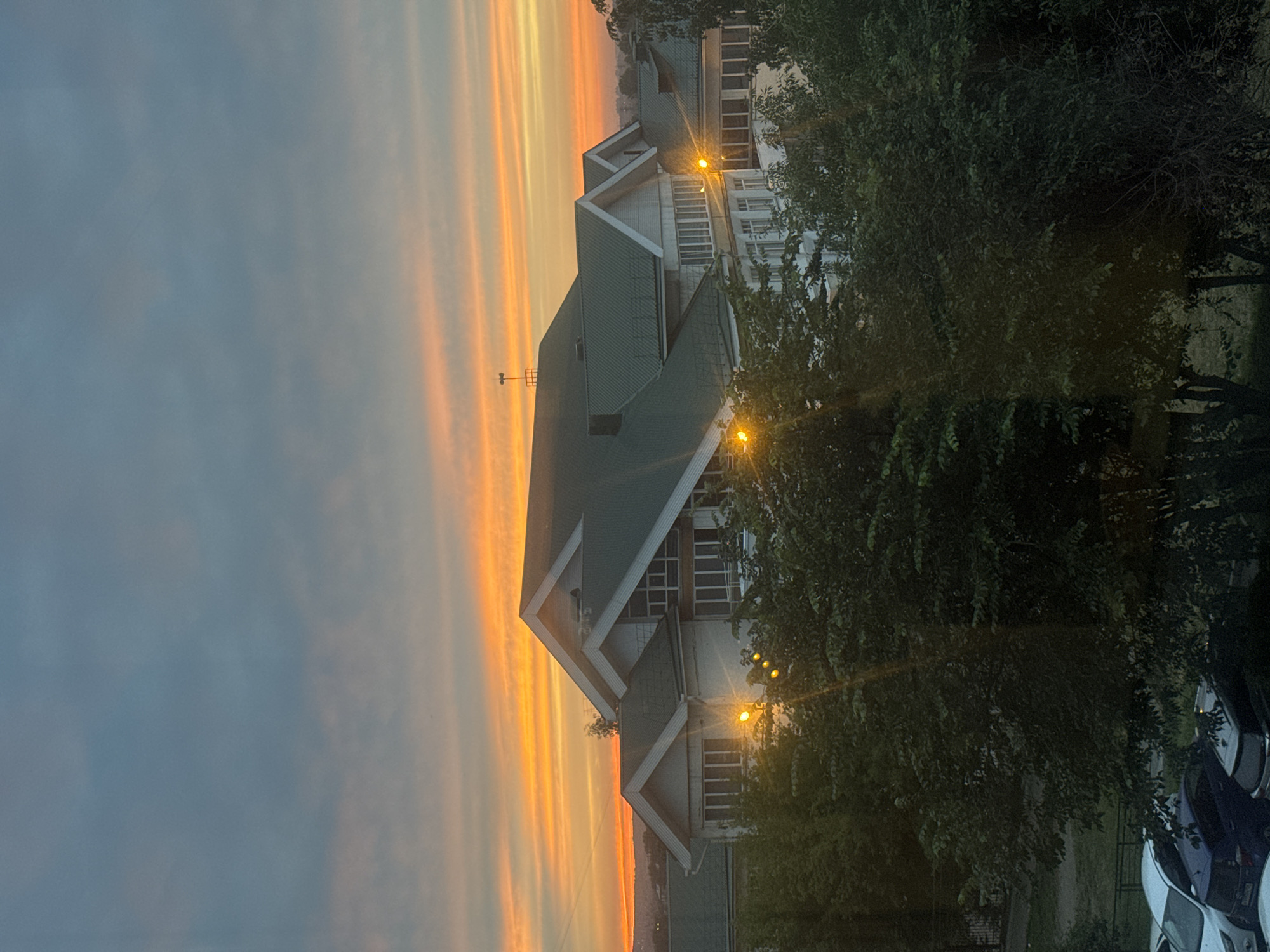 This is a vertically oriented, eye-level, medium shot of a modern building against a dramatic sunset sky. The building, with a dark green, multi-pitched roof, has a white facade and several large windows. It appears to be a residential or institutional building, possibly an apartment complex or a school. Exterior lights are visible on the facade, casting a warm glow. In the foreground, lush green trees obscure the lower portion of the building, with their leaves forming a dense canopy. Below the trees, the roofs of several cars are visible, suggesting a parking area. The sky above is a vibrant mix of orange, yellow, and blue, indicating the sun is setting. The overall impression is one of urban twilight, with nature and man-made structures coexisting. The scene is likely located in Irkutsk, Russia, based on the metadata. There are no people or animals visible in the frame.