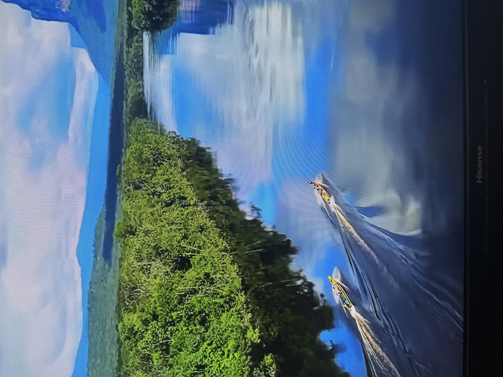 The image displays a wide, calm river with two narrow, yellow boats traveling downstream, leaving wakes behind them. The river's surface reflects the bright blue sky and scattered white clouds, as well as the dense, green foliage of trees lining the left bank. In the background, rolling hills transition into hazy blue mountains under a clear sky with some cloud formations. The scene is likely a natural landscape, possibly a tropical or subtropical region, given the lush greenery and the style of the boats. There is no indication or visual element that connects this image to the city of Kyiv, Ukraine. The bottom of the frame shows the word "Hisense" printed on what appears to be a television screen or monitor.