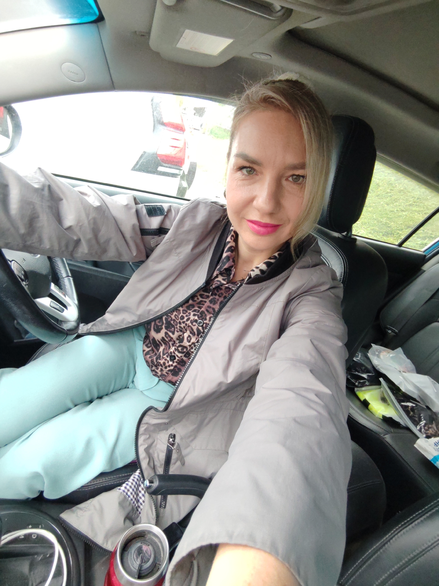 A woman is sitting in the driver's seat of a car, taking a selfie. She has blonde hair and is wearing bright pink lipstick, a leopard-print top, a gray jacket, and light blue pants. The car's interior is visible, including the steering wheel, dashboard, and seats. Outside the car, other vehicles and some greenery can be seen.