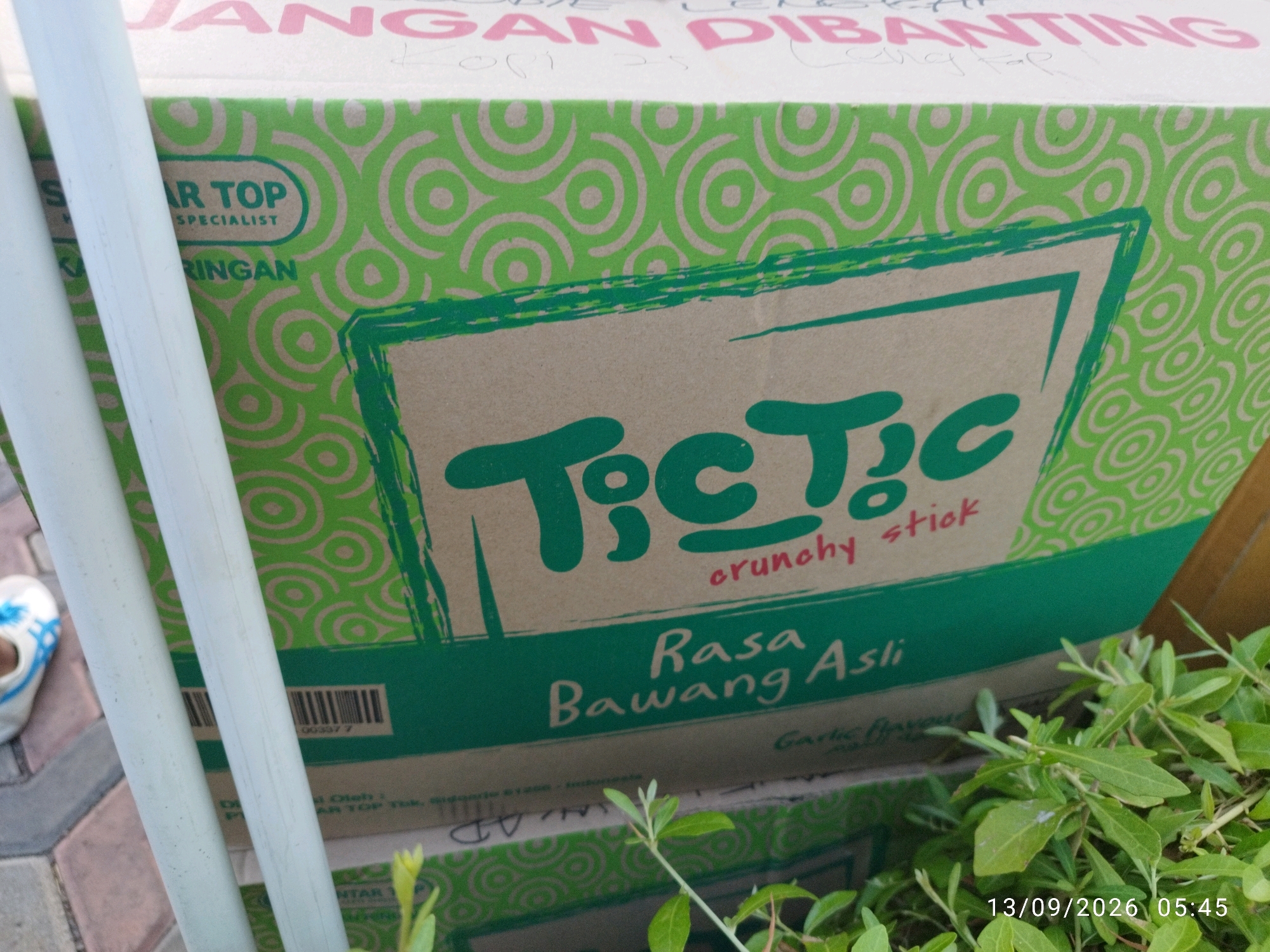 This image shows a close-up view of a green and tan cardboard box, likely containing food products, with a vibrant green circular pattern. The text on the box is in Indonesian. Prominently displayed is the logo "Tic Tac" in a handwritten font, with "crunchy stick" written below it in smaller print. Below this, "Rasa Bawang Asli" translates to "Original Onion Flavor." A barcode is visible on the left side of the box. The box appears to be outdoors, on a paved surface that resembles brickwork. Green foliage is present in the foreground and to the right, partially obscuring the lower portion of the box. In the background, two white cylindrical objects, possibly pipes or poles, are visible. The timestamp "13/09/2026 05:45" is overlaid on the lower right corner of the image. The overall impression is that of a product being stored or displayed outdoors, possibly in a market or delivery area in Mojokerto, Indonesia, though no specific landmarks pinpoint the city.