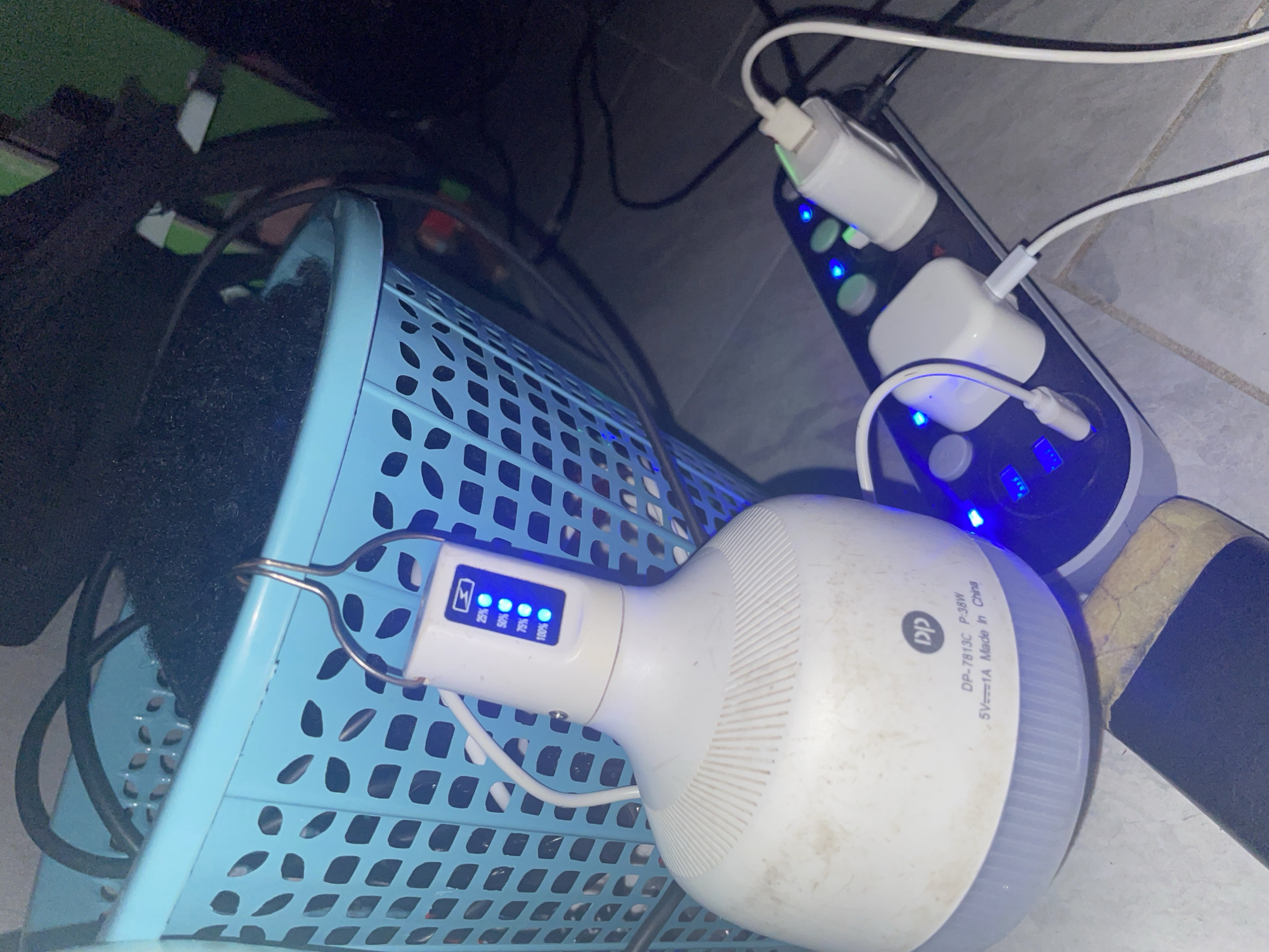 In Iyafin, Nigeria, a white LED bulb light with a hook is attached to the side of a blue plastic laundry basket filled with dark, fuzzy material. The bulb has a digital display showing battery charge levels, with lights illuminated for 25%, 50%, 75%, and 100%. The bulb is connected by a white cable to a power strip, which is plugged into an electrical outlet. The power strip has multiple sockets and several white power adapters, one of which is a white USB charger. The scene is set on a tiled floor, with cables and other objects visible in the background.