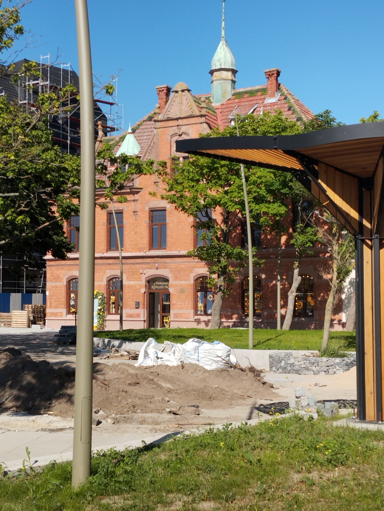 This media file shows an outdoor scene on a bright, sunny day with a clear blue sky. The primary subjects are a red brick building with a tiled roof and a prominent spire, and the surrounding area which appears to be undergoing some construction or renovation.
The building, likely a historical or municipal structure, features ornate architectural details, including arched windows and decorative gables. A small green-roofed turret is visible on one side, and a larger green spire tops the main structure. The facade has several windows, some of which display decorative elements or possibly store displays. Above one of the entrances, there is text in Cyrillic script that appears to read "Янтарное Волшебство" which translates to "Amber Magic".
The foreground and middle ground show signs of active work. Piles of dirt and sand are present, along with construction materials such as wooden pallets and large white bags, possibly containing construction supplies or soil. A modern-looking wooden and metal structure with a slanted roof stands to the right, partially obscuring the view of the building. Several trees with lush green foliage are scattered throughout the scene, casting dappled shadows on the ground. A tall, grey pole, possibly a lamppost or utility pole, stands in the foreground. The overall impression is of a public space in Zelenogradsk, Russia, transitioning or being maintained, with a mix of historical architecture and contemporary infrastructure. The sunlight suggests it is daytime.