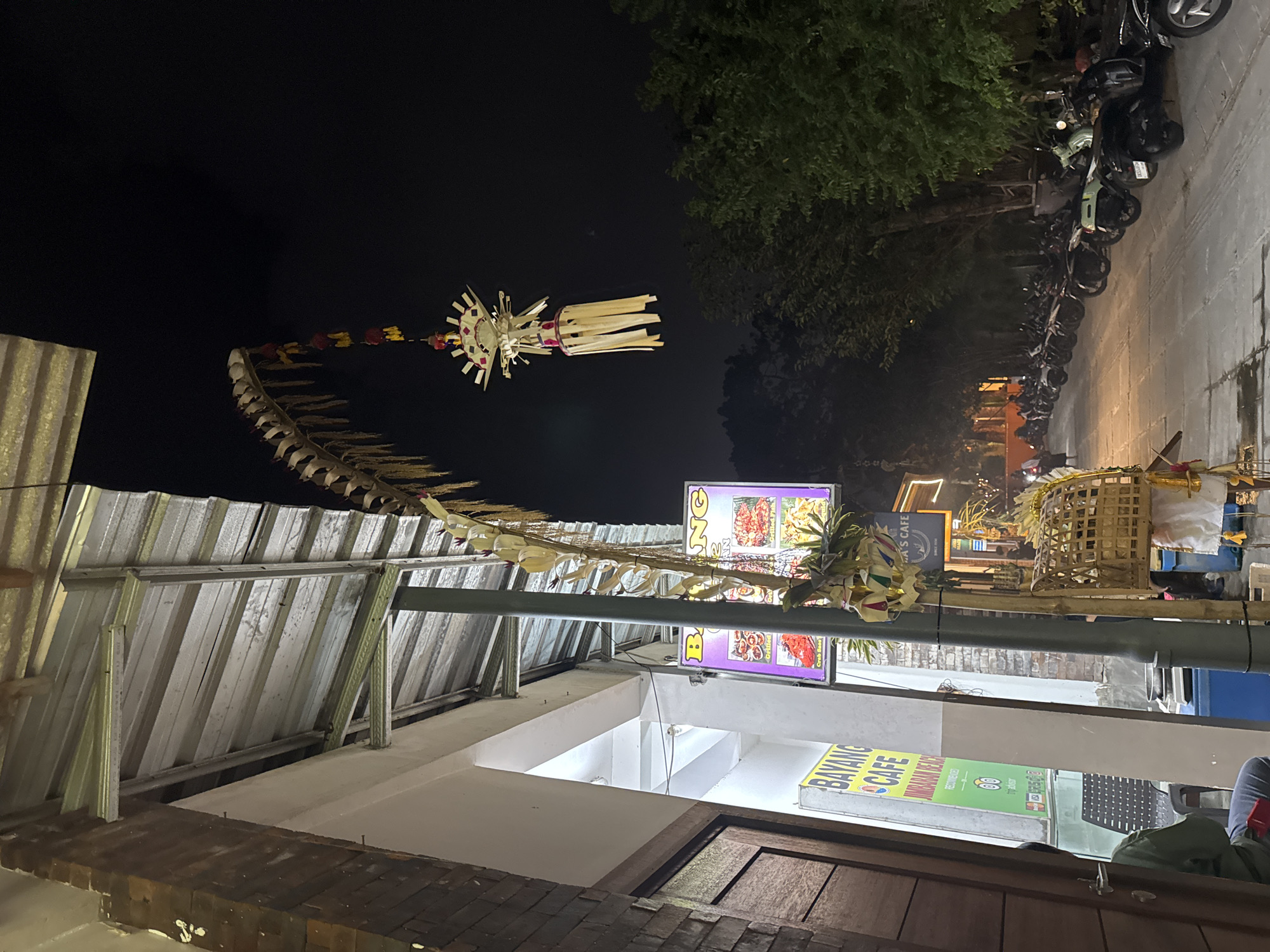The image depicts a street scene at night in Kuta Selatan, Indonesia, likely near a beachfront area given the presence of cafes and a scooter parking lot. A prominent, ornate Balinese "penjor" or decorative bamboo pole hangs from a building's awning, adorned with woven decorations and colorful tassels, extending into the dark sky. Below, a sign for "BAYANG CAFE" is visible, indicating a food establishment. A row of parked scooters and motorcycles lines the street, suggesting a popular area for visitors. The architecture features brick and metal structures, common in coastal Indonesian towns. Trees with dense foliage flank the street, creating a canopy effect. The overall atmosphere is that of a lively evening, with artificial lighting from signs and street lamps illuminating the scene.