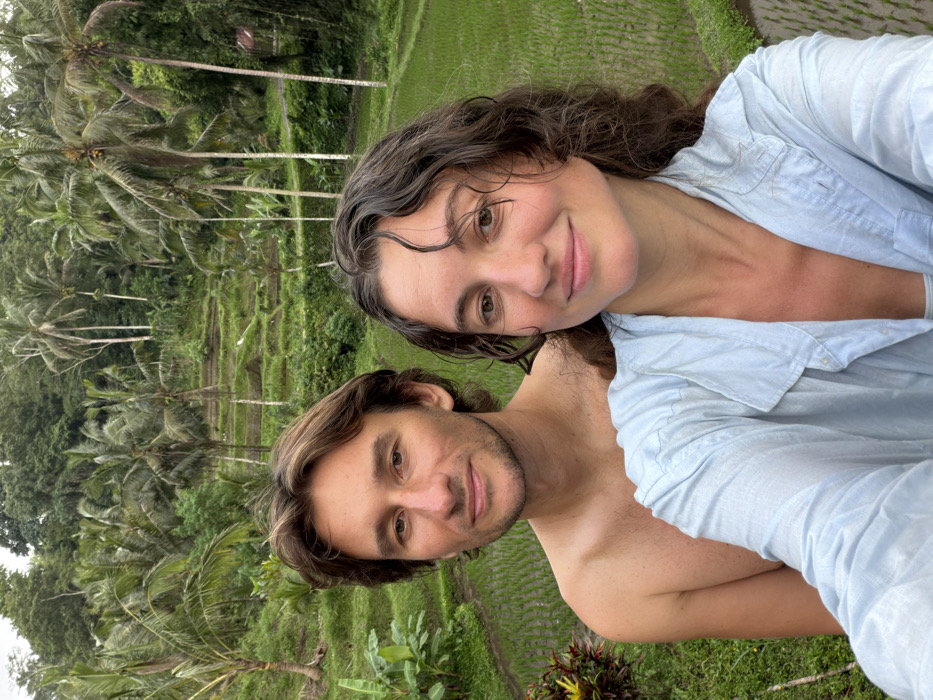 An outdoor portrait features two individuals against a backdrop of terraced rice fields and tropical vegetation in Tegal Suci, Indonesia. The female subject, positioned closer to the camera on the right, has dark, curly hair and a light complexion, wearing a light blue unbuttoned shirt over a white undergarment. She is smiling and appears to be holding the camera for a selfie. Behind and slightly to her left stands a male subject with light brown, wavy hair, short facial stubble, and a bare torso; he also faces the camera with a faint smile. The background is dominated by multiple tiers of vibrant green rice paddies, some appearing wet, interspersed with numerous tall palm trees and dense jungle foliage under an overcast sky.
