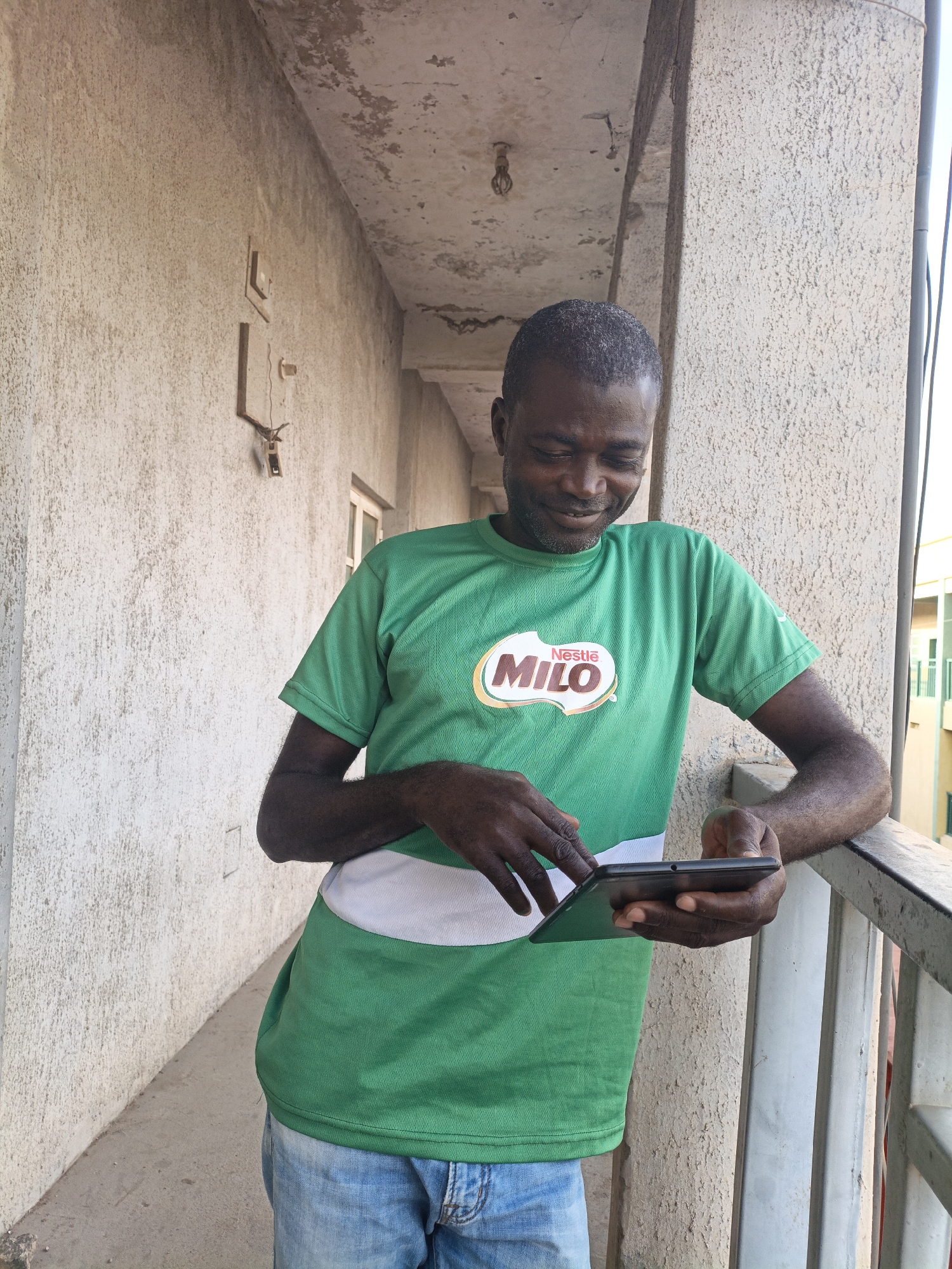 A dark-skinned man with a shaved head and a beard, wearing a green t-shirt with a white stripe across the chest and a "Nestle Milo" logo, and blue jeans, stands on a covered walkway or balcony. He is looking down at a black tablet device he is holding in his hands and appears to be smiling. The walkway has a rough, light-colored textured wall on the left, a concrete pillar on the right, and a metal railing. The ceiling of the walkway is concrete and appears weathered, with exposed conduits and a light fixture. In the background, a building with windows is visible. The scene is located in Fagge C, Nigeria.