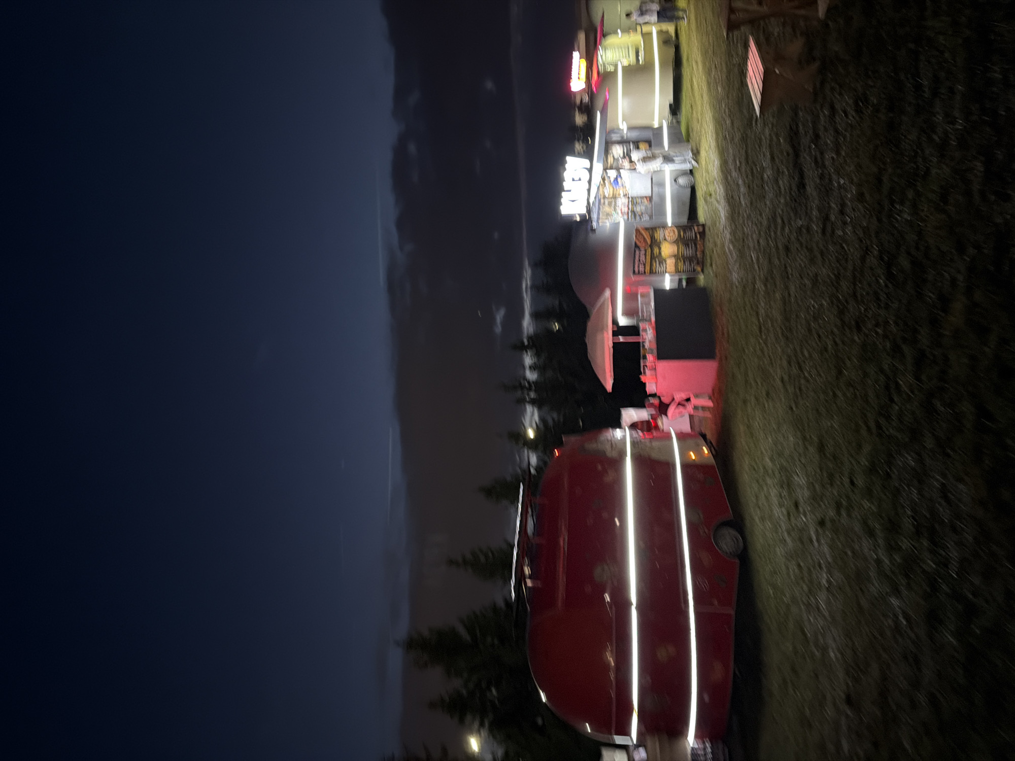The image depicts an outdoor scene at night, likely a food truck or festival setting. In the foreground, a large, red, rounded object, possibly a food trailer or a vehicle, is illuminated by vertical white LED lights. A black, boxy object, possibly a counter or a table, is situated in front of the trailer. To the right of the trailer, a grassy area is visible.
Further back, several food stalls are illuminated by bright lights. One stall features neon signage that reads "KITCHEN" in large white letters. Another stall appears to have a menu board displaying images of food items. People are visible in the background, suggesting activity and customers. The overall ambiance is lively and suggests a nighttime gathering or event. The sky is dark, indicating it is nighttime, and the ground appears to be slightly wet, hinting at recent or ongoing rain.