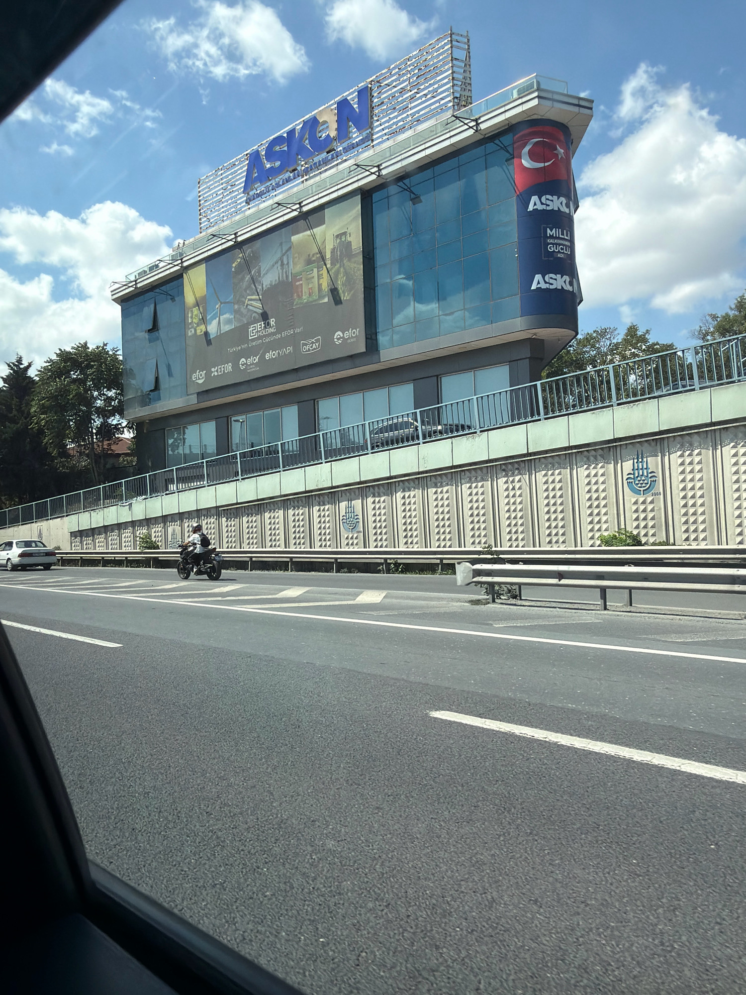 The image shows a street-level view of the ASKON building in Zeytinburnu, Turkey. The building features a prominent "ASKON" sign at the top, with a large banner displaying advertisements on its facade. Below the building, a highway is visible with a white car and a motorcyclist traveling on it. The highway is flanked by a retaining wall decorated with a repeating geometric pattern and a stylized emblem of a hand with three wavy lines above it, which is the symbol of Istanbul. The sky is blue with scattered clouds.