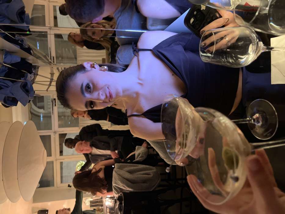 A woman in a black top looks at the camera. She has brown hair pulled back and wears hoop earrings. The image seems to be taken in a restaurant or bar, with multiple people and a lighting fixture visible in the background. Several wine glasses are in the foreground. The scene might be captured from a reflective surface, creating a sense of depth and repetition.