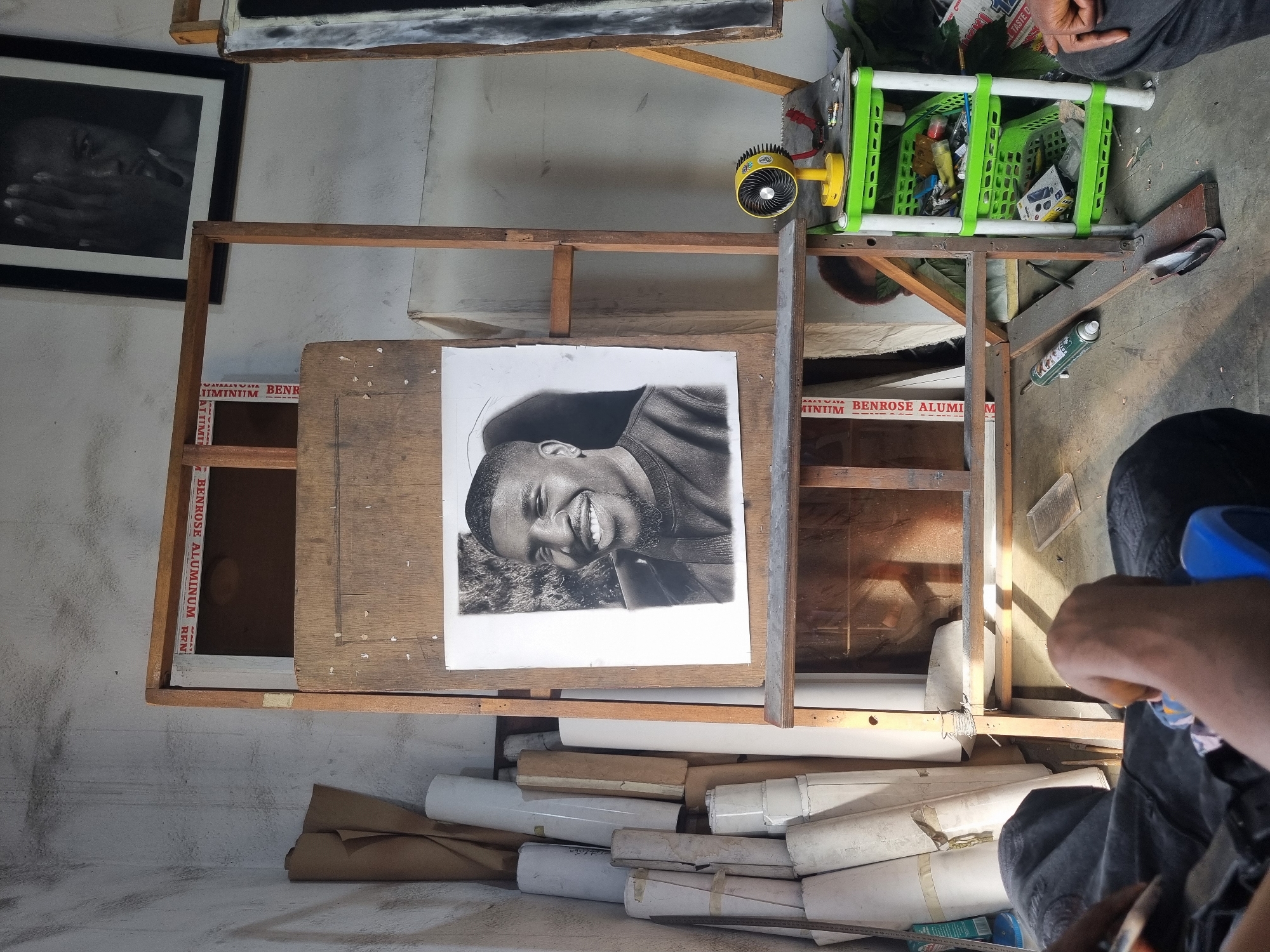 The image shows a cluttered workspace in Onitsha, Nigeria. At the center, there's a wooden easel holding a charcoal portrait of a smiling man. To the left, a framed black and white picture hangs on a stained wall. A small yellow fan is mounted above the portrait. A green plastic basket filled with items is visible on the top right of the frame. Numerous rolled paper documents are stacked beneath the easel. A hand holding an object is visible on the right bottom corner. The workspace appears to be in a state of disarray.