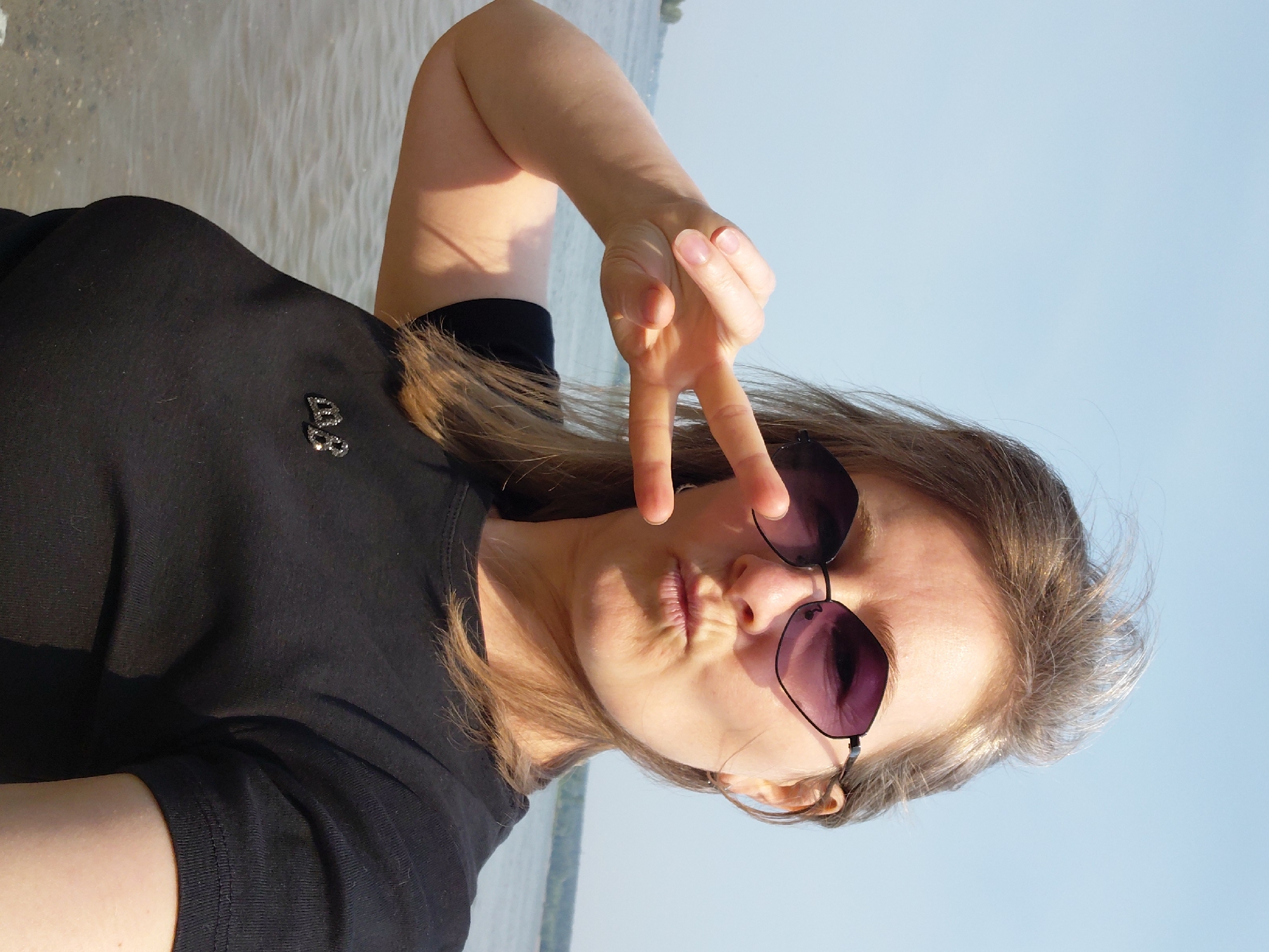 A woman is taking a selfie outdoors near a body of water, possibly the Ob River, given the context of Novosibirsk, Russia. She has medium-length, light brown hair and is wearing sunglasses with purple lenses and a black t-shirt. She is making a peace sign with her left hand and puckering her lips. The background shows a calm body of water with a distant tree line under a clear, light blue sky. A small, metallic badge with the letters "AB" is visible on her t-shirt. The lighting suggests it is daytime.