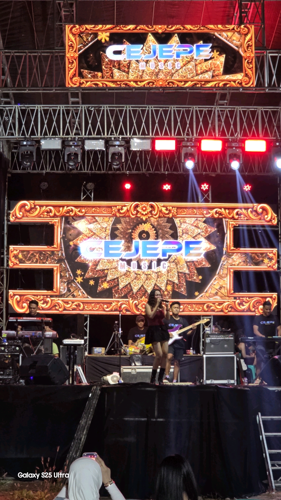 The image shows an outdoor stage setup for a music performance in Jaken, Indonesia. A large LED screen displaying the text "CEJEPE MUSIC" with intricate golden ornamental borders dominates the background. Below the screen, a band is performing on a black stage. A female singer in a red top and black skirt is in the center, holding a microphone. To her left, a keyboardist is playing an instrument. To her right, a guitarist is playing an electric guitar, and another musician is visible behind him. Several other band members and equipment, including speakers and more keyboards, are also present on the stage. The lighting includes bright spotlights and red stage lights. In the foreground, a person in a white hijab is holding up a phone to record the performance. The overall environment suggests a nighttime event.