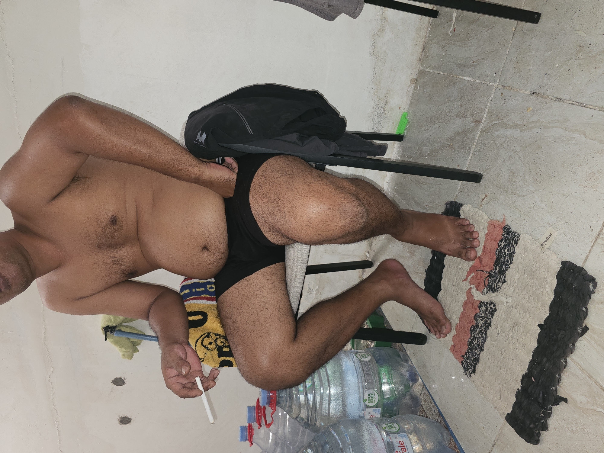 The image shows a shirtless man with dark hair sitting on a chair, holding a cigarette. He is wearing black underwear and his legs are exposed. There are several large water bottles behind him, some with labels indicating they are BPA-free. A black jacket is draped over the back of the chair. A colorful rug is on the floor in front of the man, and his bare feet are resting on it. The background is a light-colored wall with some discoloration and two circular holes. The floor appears to be tiled. The overall scene suggests an indoor or semi-outdoor setting, possibly in a utilitarian or storage area.