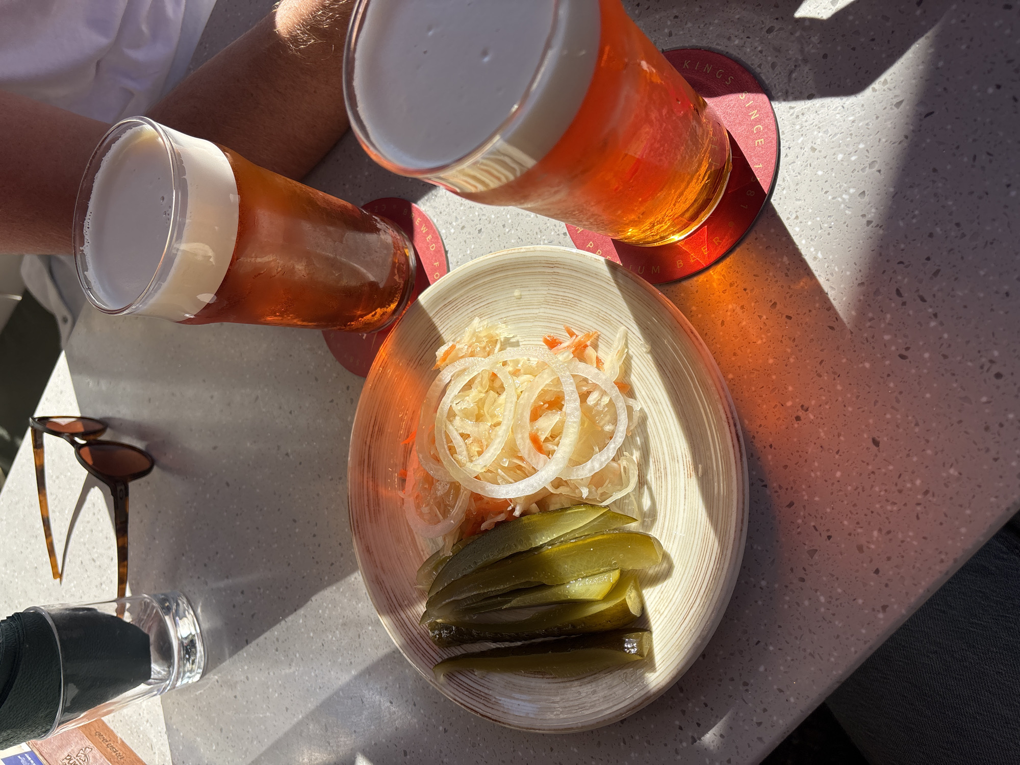 In this overhead shot, two glasses of amber beer with frothy white heads sit on a textured countertop. One glass is in the foreground, tilted slightly, revealing its contents. The other glass, further back and angled, casts a warm orange glow on a red circular coaster that reads "KINGS SINCE 1887". Next to the glasses is a small, light-colored bowl containing sauerkraut and dill pickle spears. The sauerkraut is piled high, topped with thin slices of white onion. The pickles are a deep green, cut lengthwise, and layered in the bowl.
The scene appears to be outdoors, suggested by the strong sunlight casting sharp shadows across the table and on the beer. The light indicates it's likely daytime, possibly mid-morning or afternoon, given the angle of the sun. A pair of tortoiseshell sunglasses rests on the table near a clear glass that holds a dark object, possibly a napkin. The countertop itself has a speckled pattern with flecks of grey, white, and possibly pink.
The presence of beer and pickles suggests a casual setting, perhaps a beer garden or an outdoor cafe. The overall impression is one of relaxation and enjoyment, bathed in sunlight. While the image is primarily focused on food and drink, the suggestion of people's arms and clothing at the edges hints at a social gathering. The context of Saint Petersburg, Russia, doesn't introduce any specific visual cues within the image itself but provides a geographical frame.