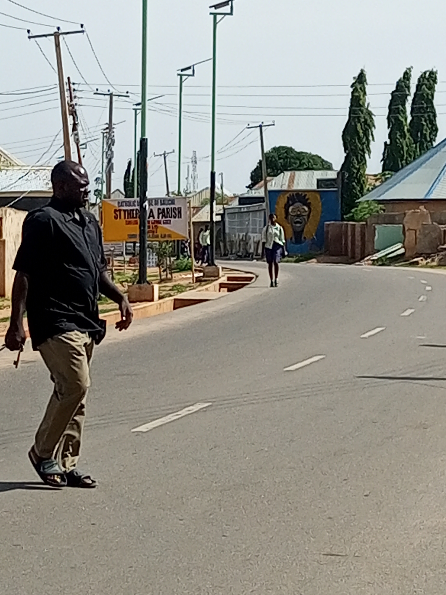 A middle-aged African man wearing a black short-sleeved shirt and beige pants is walking across a paved road in Gombe, Nigeria. He is carrying a set of keys in his right hand. In the background, a woman in a light-colored shirt and dark skirt is walking away from the camera on the sidewalk. To the right of the woman is a mural of a person's face with a yellow halo. Several utility poles with electrical wires are visible throughout the scene. The road has white dashed lines marking the lanes. The weather appears to be clear and sunny.