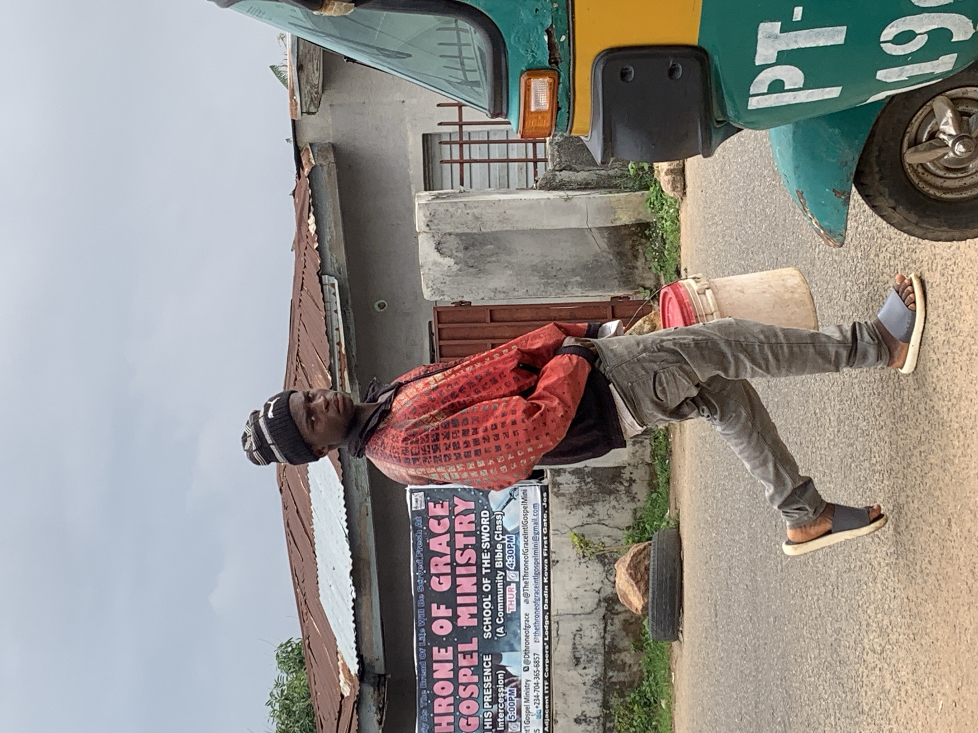 A medium shot depicts a person standing on a paved road next to a three-wheeled vehicle, potentially an auto-rickshaw, with a container or bucket resting on the vehicle. The person is wearing a knitted cap, a patterned red jacket, grey trousers, and sandals. A sign for "Throne of Grace Gospel Ministry" is affixed to a building or wall in the background, indicating the location as being near the ITF Corpers' Lodge, Dadin Kowa First Gate, Jos, Nigeria. The architecture suggests an urban or semi-urban setting in Nigeria.