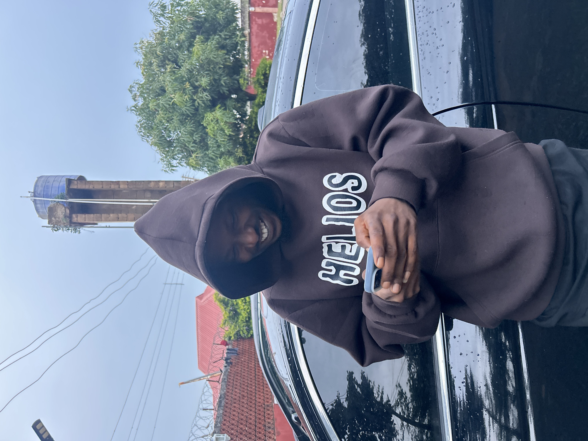 A young Black man wearing a dark brown hooded sweatshirt with white lettering that reads "HELIOS" is standing outdoors next to a black car. He is looking down at his phone, which he is holding with both hands. The man has a wide smile on his face. In the background, there is a tall, weathered water tower with a blue tank at the top. Behind the water tower, there is a large green tree. To the left of the water tower, there is a red-roofed building and a fence with barbed wire. The ground appears to be wet, possibly from rain, with visible water droplets on the car's surface. The sky is overcast and light blue. The scene is consistent with an outdoor location in Jos, Nigeria.