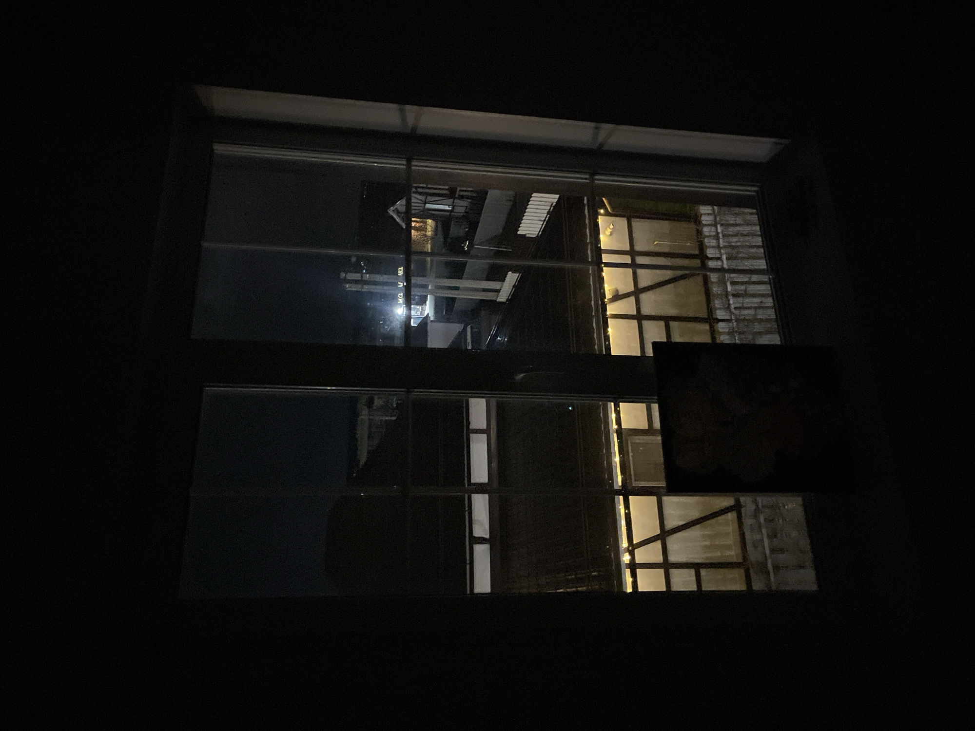 A view from a dark room through a window shows an outdoor scene at night. The window is divided into panes. Reflections are visible on the glass. Outside, there's a building with illuminated windows. The location cannot be determined from the provided image.