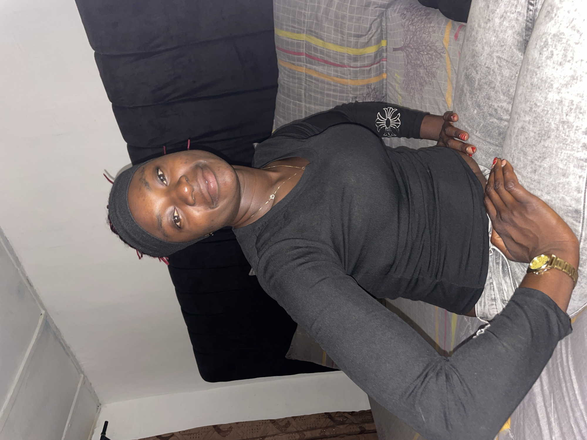 A dark-skinned adult female, appearing to be in her 20s or 30s, is seated on a bed in an indoor setting in Jos, Nigeria. She looks directly at the camera with a slight smile. Her hair, possibly dreadlocks or braids with some red strands, is partially covered by a black headband. She wears a black long-sleeved V-neck top featuring a white symbolic design on the left forearm and light grey acid-wash jeans. A gold-colored chain necklace with a small charm is visible around her neck, and a gold-colored wristwatch is on her left wrist. Her right hand, resting on her lap, displays red nail polish with white and gold accents on several fingers. She is positioned against a dark, vertically tufted headboard. The bed is covered with a grey duvet featuring patterned stripes in yellow, pink, and red, over a subtle checkered pattern. A plain white wall is visible behind her, with a brown patterned curtain partially seen on the far left. The white ceiling above shows panel seams and some minor discoloration in the corner. Artificial lighting illuminates the scene.