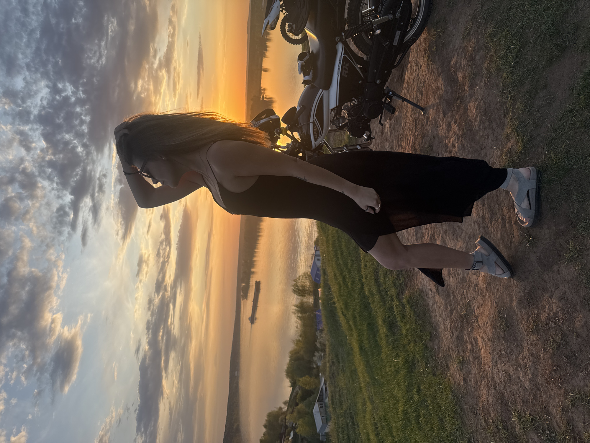 A woman stands near a motorcycle on the bank of a river in Olkhovka, Russia. The woman has long hair and wears a black dress and sandals. She has one hand touching her head. A motorcycle sits to her right. The river and sunset are in the background. Several buildings are on the bank of the river. Barges float on the river.