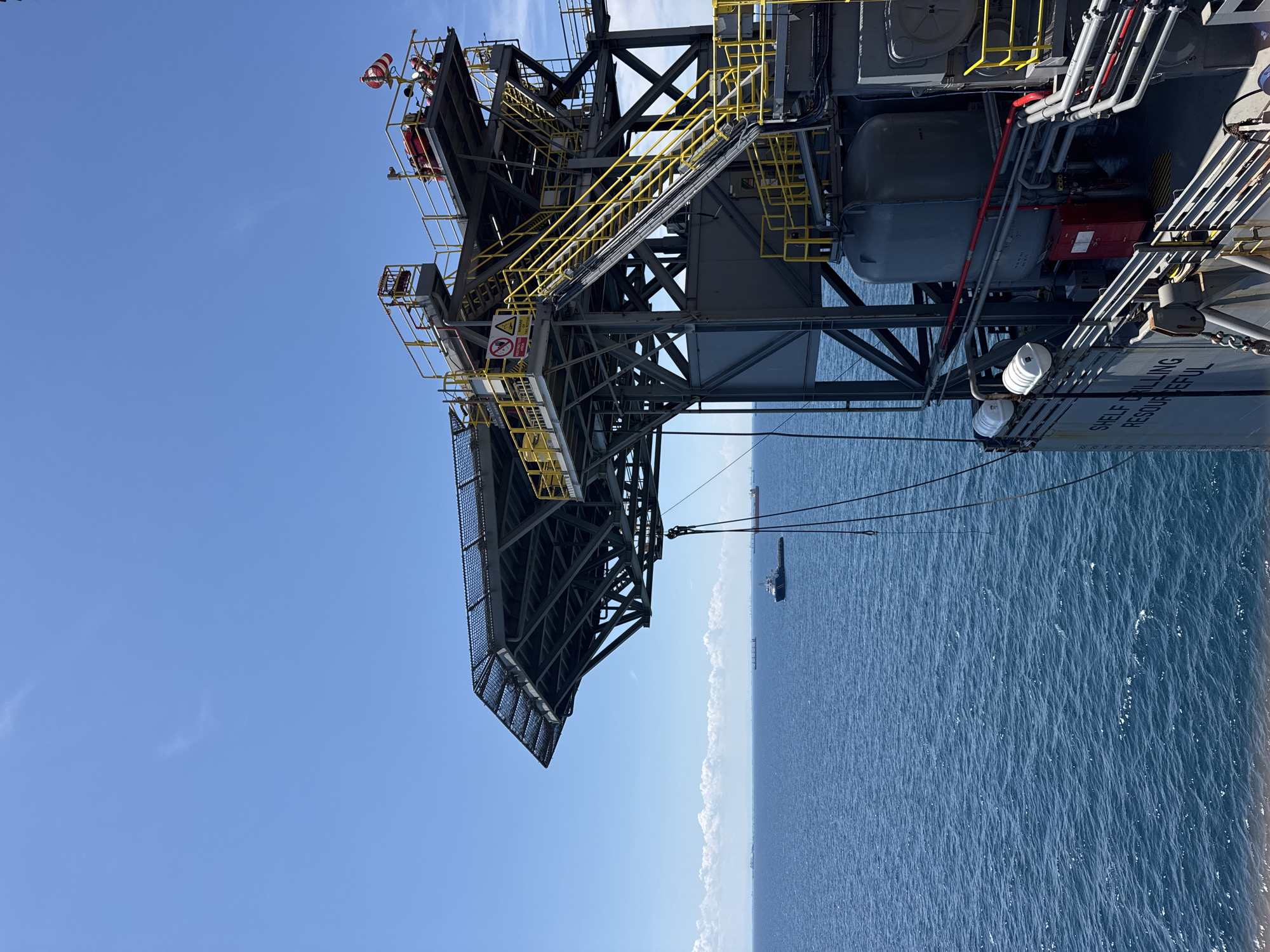 An offshore drilling platform helideck and structure are visible against a clear blue sky and the ocean. Several ships are scattered on the water in the distance, with a small tugboat positioned closer to the platform. The helideck has a yellow railing and safety equipment, including life rafts and a windsock. The main structure of the platform features yellow staircases and industrial pipes. The text "SHELF DRILLING RESOURCEFUL" is visible on the side of the platform. The location is not in a city in Italy, but rather on the open sea.