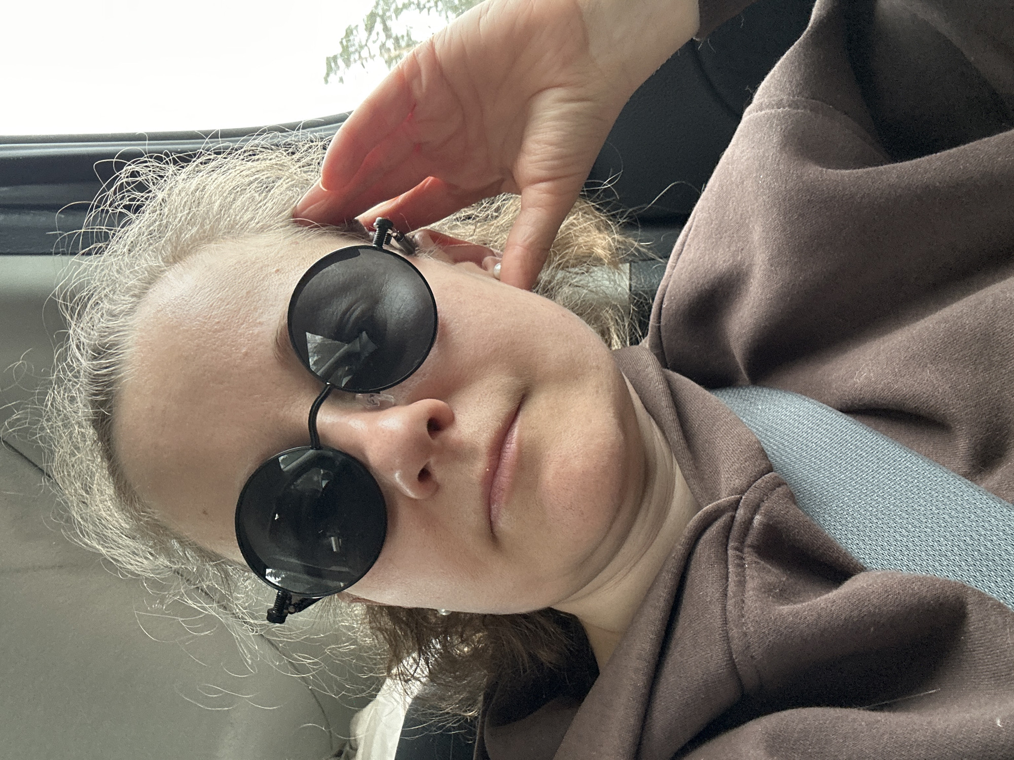 The image depicts an adult individual in the city of Minino, Russia, situated inside a vehicle. The subject is female, facing the camera, wearing dark, round-framed sunglasses. Her light, curly hair shows visible grey strands. A small white blemish is present on her upper lip. She is attired in a brown hooded sweatshirt. A grey fabric seatbelt traverses diagonally across her chest and shoulder. Her right hand rests near her right temple. A pearl-like stud earring is visible in her right earlobe. The background reveals the vehicle's light-colored headliner, a dark window frame, and a blurred exterior scene containing foliage and sky.