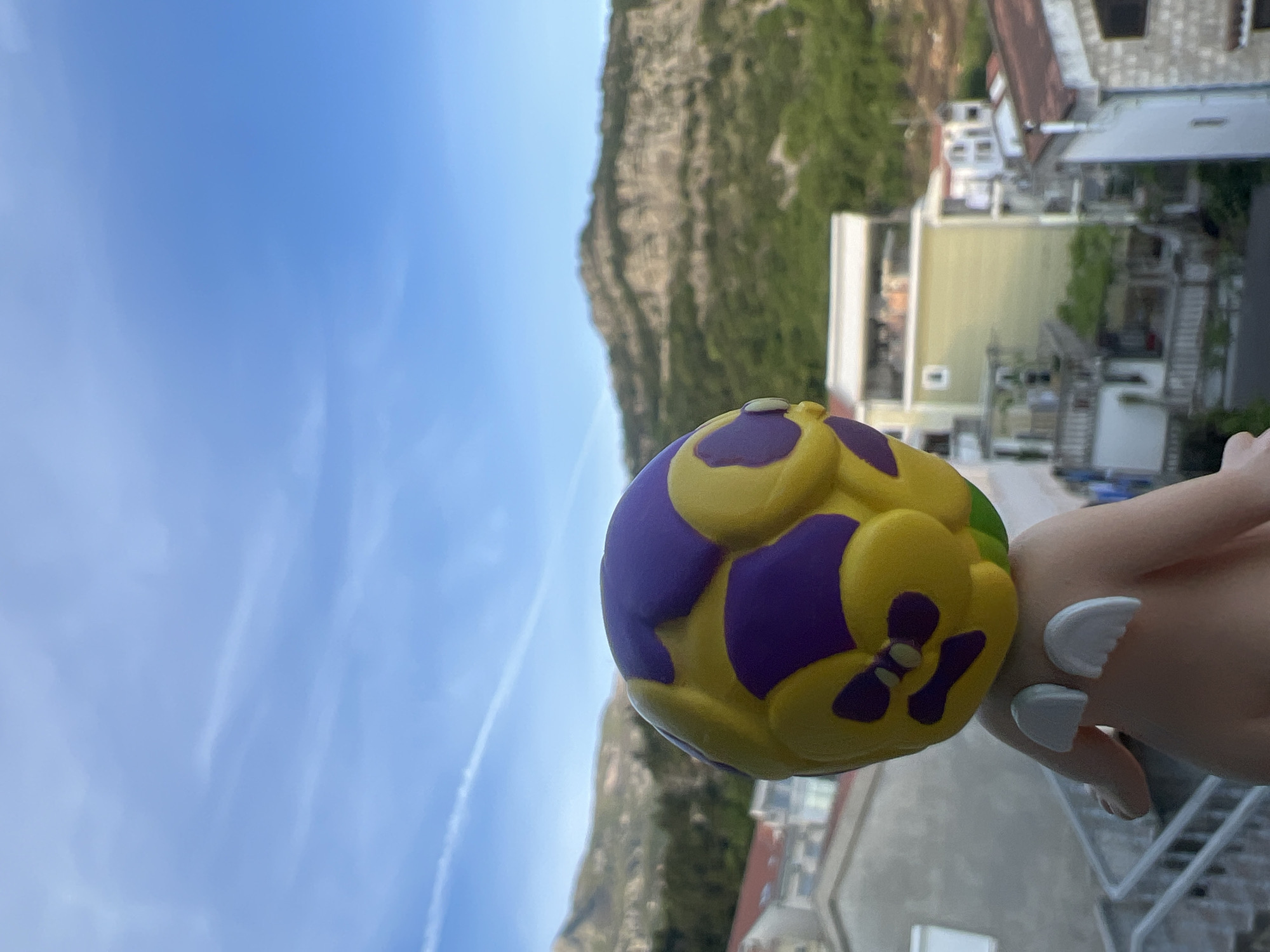 The image shows a hand holding a yellow and purple flower-shaped stress ball against a backdrop of Budva, Montenegro. The sky is blue with some visible contrails. In the background, there's a hillside covered in greenery, along with buildings characteristic of the city, including residential houses with light-colored facades and red roofs. The buildings are terraced down the hillside. The hand holding the stress ball has pale skin and visible white nail polish.