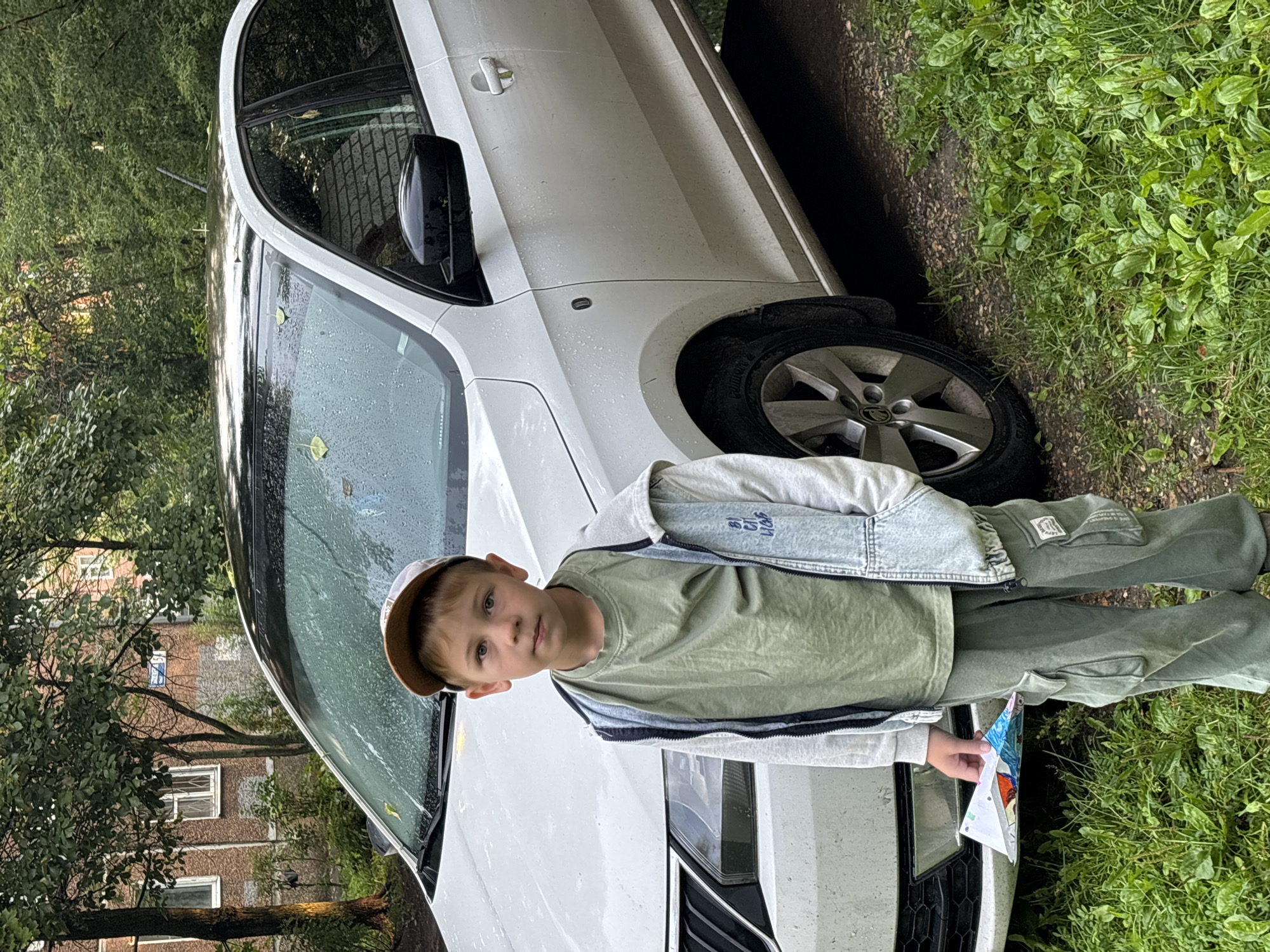 In the city of Rusanovo, Russia, a young boy stands in front of a white car parked on a gravel surface next to a grassy area. The car's windshield is wet, indicating recent rain, and a few leaves are scattered on its surface. The boy is wearing a green t-shirt, a light-colored hooded jacket, and green cargo pants. He has a brown and beige baseball cap on his head. In his left hand, he holds a folded piece of paper with colorful drawings on it. The background features trees and a brick building with windows. The scene appears to be an outdoor setting, possibly a residential area.