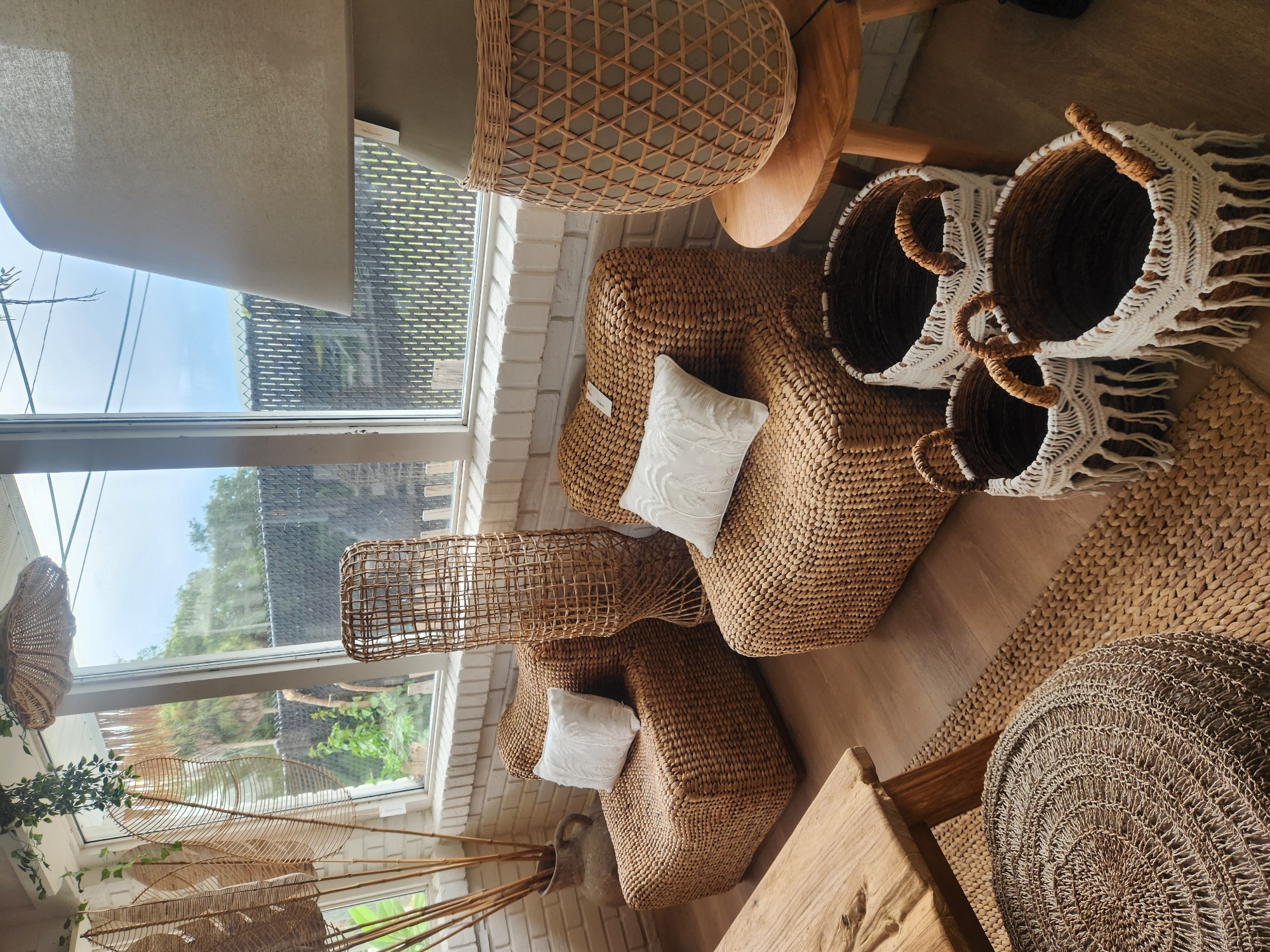 This is a forensic, matter-of-fact description of a store interior in Maitland, United States of America. The image displays a collection of natural, woven furniture and decor items. In the foreground, a wooden coffee table with a rough-hewn top sits on a woven rug. To the right of the table, three decorative woven baskets with white fringe and brown handles are arranged on the floor. A round, woven ottoman or pouf is positioned to the left of the coffee table, also on a rug.
Further into the scene, two cube-shaped wicker armchairs are placed side-by-side. Each armchair has a white decorative pillow on its seat. Behind the armchairs, a tall, cylindrical woven floor lamp or decorative accent stands to the left. To the right of the armchairs, a large floor lamp with a cream-colored shade sits on a wooden side table. The background features a large window that looks out onto an outdoor area with greenery and a fence. The walls of the interior space are constructed of white brick. The overall aesthetic suggests a bohemian or coastal-inspired retail environment. There are no people visible in the image.