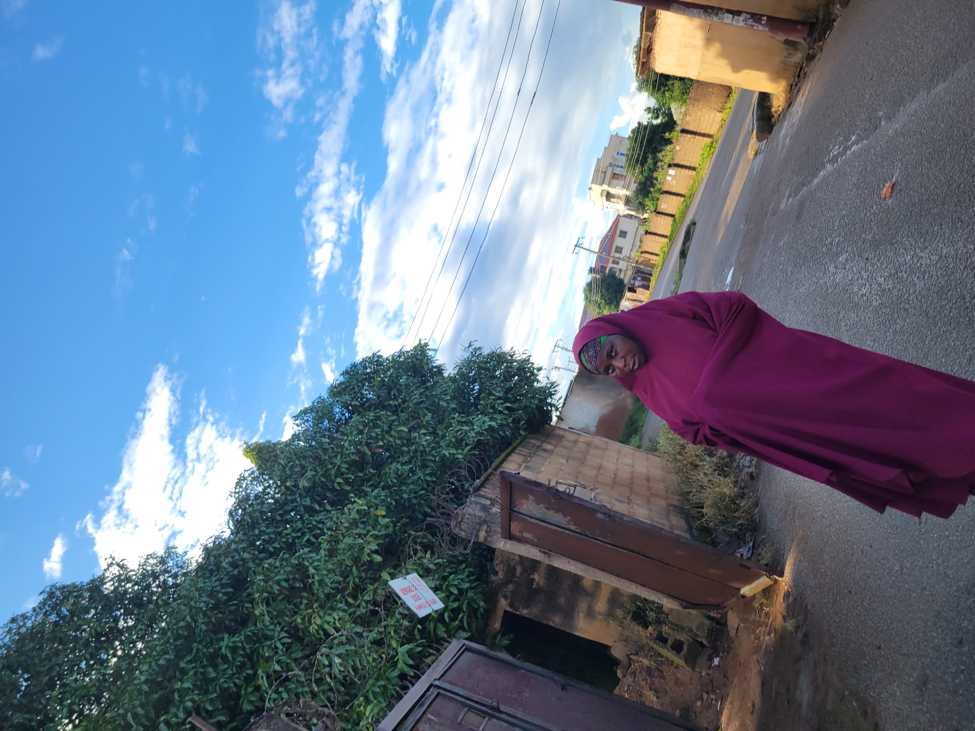 A young Black woman wearing a magenta hijab and a matching magenta garment is standing on an asphalt road. She is facing the camera and looking directly at it. The background includes a tree with dark green leaves, a rustic metal gate and door, a brick wall with barbed wire on top, and several houses in the distance. The sky is blue with scattered white clouds. The overall impression is of a casual outdoor scene in a residential area.