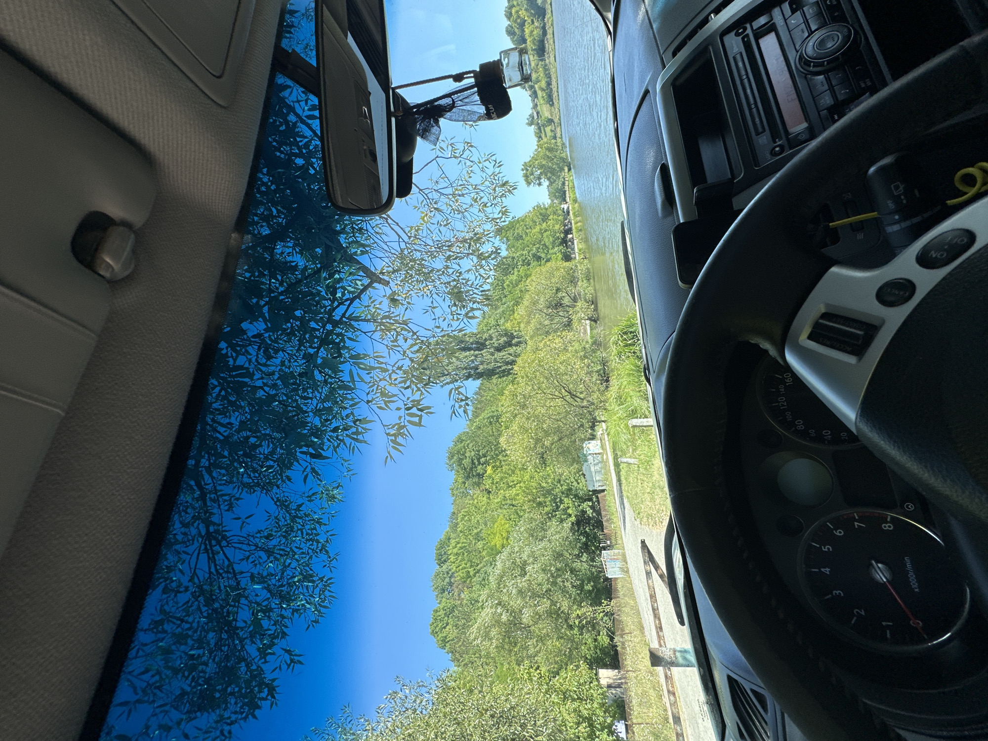 The image is taken from inside a car, looking out through the windshield. The car's steering wheel and dashboard are visible in the foreground. The view outside shows a body of water, likely a river or lake, bordered by lush green trees and hills. The sky is clear and blue. There are some structures visible along the bank of the water, and a road or path leading into the trees. The scene appears to be a serene, natural landscape. There are no people visible in the image.