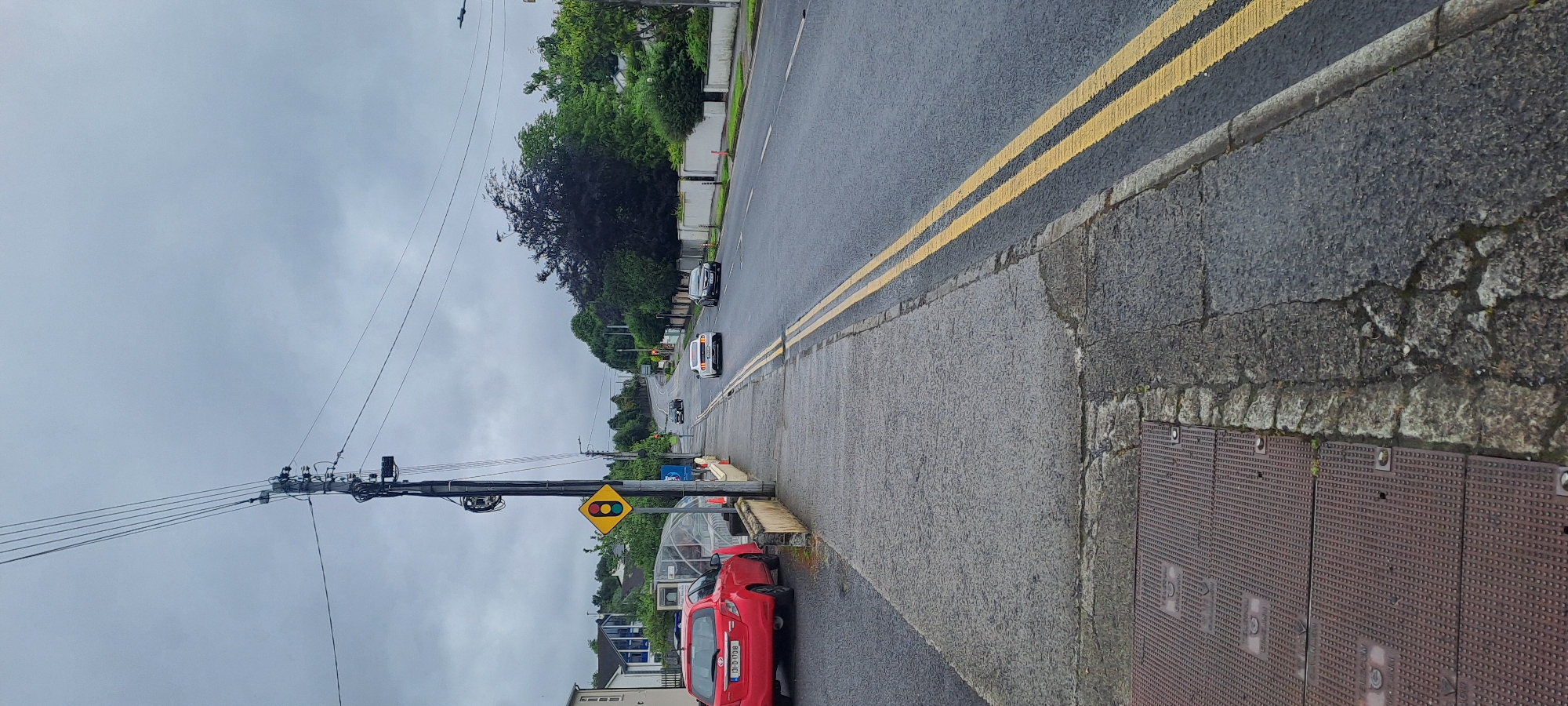 The image captures a view of a street in Castlebar, Ireland, under a cloudy sky. The street is paved with asphalt and marked with double yellow lines indicating no overtaking. A sidewalk runs along the left side of the road, bordered by a low wall. Several vehicles are visible on the road, including a red hatchback parked on the left, a white sports car and a black sedan driving away from the camera, and two other cars further in the distance. A traffic light is positioned on the sidewalk near the red car. Power lines and poles are prominent features in the sky and along the street. The surrounding environment includes houses and lush green trees. The sidewalk has several metal manhole covers. The overall impression is of a suburban street scene on an overcast day.