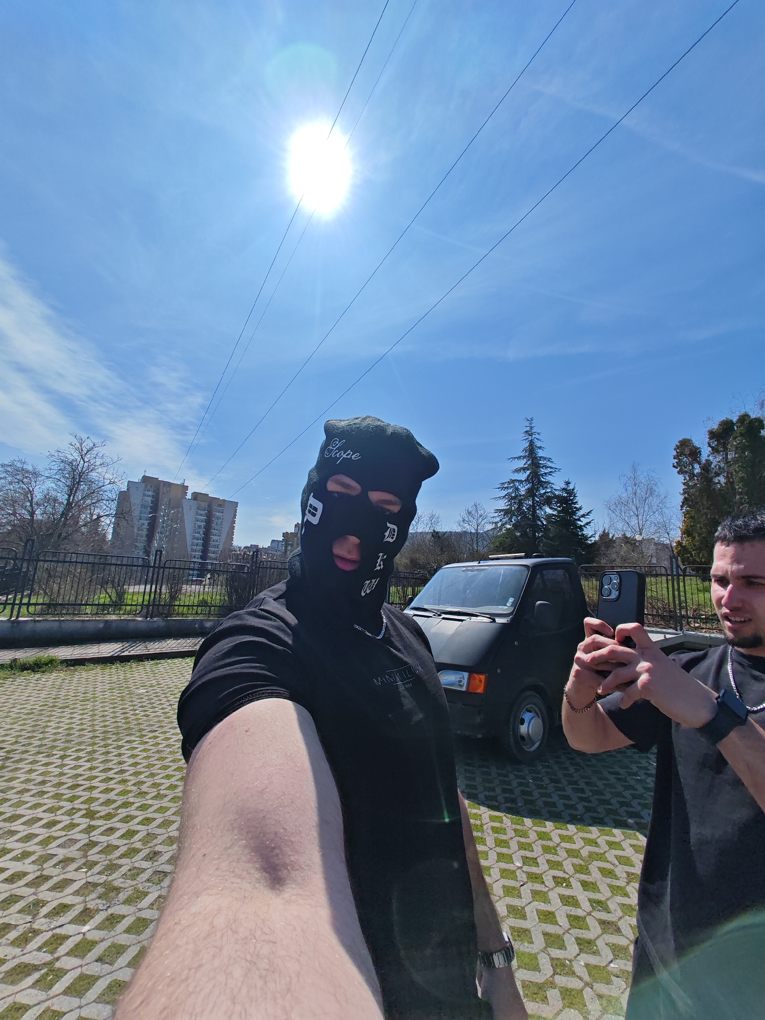 The image depicts an outdoor daytime scene in Shumen, Bulgaria, specifically within a residential area identifiable by multi-story apartment buildings in the background, possibly near ul. "Preslav" 20.
In the foreground, a male subject is taking a selfie. He wears a black balaclava with white lettering (including "Scope"), a black short-sleeved t-shirt, a silver chain, and a silver watch on his left wrist. His fair-skinned, hairy right arm is extended, holding the camera. To his right, a second male subject is visible from the chest up, holding a dark smartphone with a triple-lens camera horizontally with both hands, appearing to record or photograph. He wears a dark short-sleeved t-shirt, a silver chain, and a black smartwatch on his left wrist. His mouth is slightly open.
The ground beneath the subjects consists of grey interlocking concrete pavers with patches of green grass growing in the interstices. A dark-colored mini-truck or utility van is parked behind the two individuals, partially visible between them.
Behind a dark metal fence, to the left, are leafless deciduous trees and a grassy area. In the far background, several multi-story residential apartment buildings with light-colored facades and distinct architectural features (such as staggered balconies) are prominent under bright sunlight. To the right of the parked vehicle, an evergreen tree and other trees with sparse foliage are visible beyond the fence.
The sky is a bright, clear blue with faint wispy clouds. A prominent, intense sun is positioned in the upper center, generating a strong lens flare. Multiple thin, dark overhead power lines or cables traverse the upper portion of the frame diagonally from left to right.