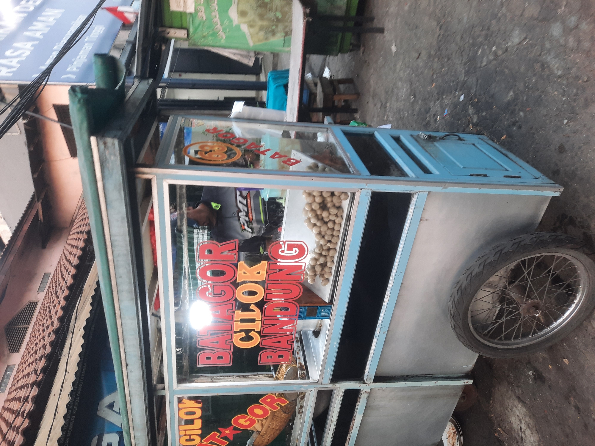 A food cart selling "Batagor" and "Cilok Bandung" is parked on a street in North Jakarta, Indonesia. The cart is made of metal and wood, painted blue and silver, and features large glass windows displaying the food and menu items. A vendor, wearing a grey shirt with "PMI" on it, is partially visible behind the cart. Piles of meatballs are visible inside the cart. In the background, buildings with tiled roofs and wires are seen, along with other street vendors and tables. The ground is paved and appears somewhat worn.