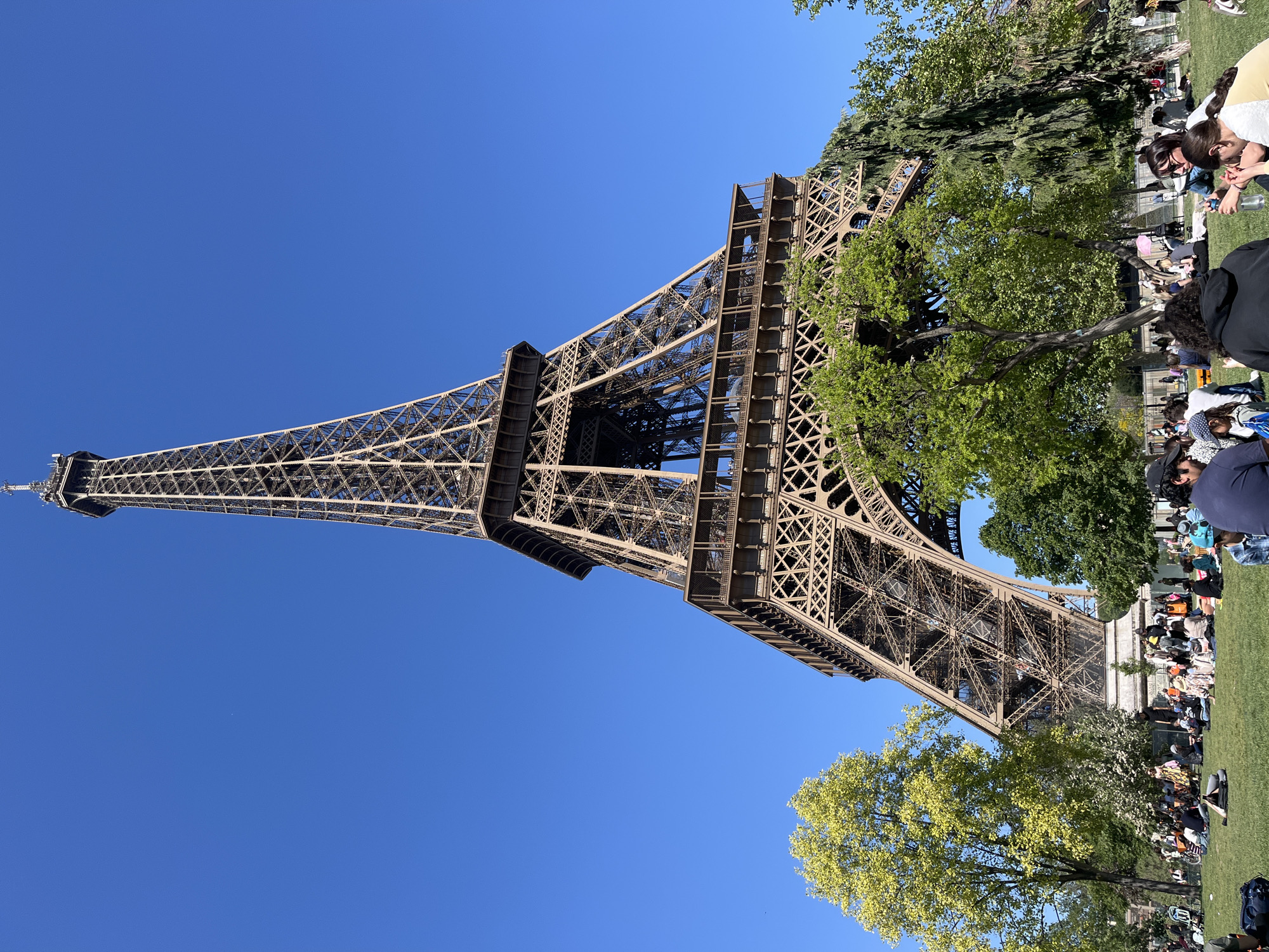 This image captures a vibrant outdoor scene featuring the iconic Eiffel Tower in Paris, France. The Eiffel Tower dominates the left and central portion of the frame, showcasing its intricate lattice structure and characteristic brown-grey color against a clear, bright blue sky. The photograph is oriented with a 90-degree clockwise rotation, causing the tower to appear to lean diagonally from the lower left towards the upper right of the frame, with its base rooted in the lower right.
In the foreground and midground, a large expanse of verdant green grass is dotted with numerous people. Individuals and small groups are seen sitting and lounging on the grass, engaged in leisurely activities such as relaxing, conversing, and possibly picnicking. Several lush green trees are scattered throughout the scene, some partially obscuring parts of the Eiffel Tower or providing shade for the people below.
The bright, unfiltered sunlight and the cloudless sky indicate a clear and pleasant day, likely mid-morning or afternoon, ideal for outdoor enjoyment. The overall atmosphere is one of relaxation and enjoyment, with people appearing to take in the views and the good weather around the famous landmark. No discernible text is visible within the image.
