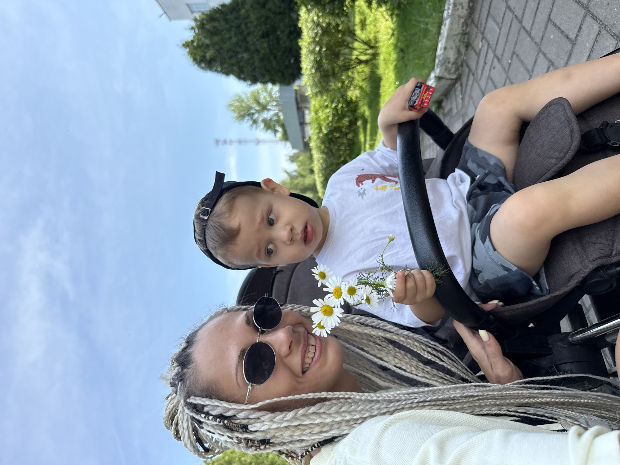 In Rusanovo, Russia, a woman with blonde braids and sunglasses smiles while holding a small bouquet of daisies near her face. Next to her, a young child in a stroller wears a white t-shirt and a black baseball cap backwards. The child is looking away from the camera. The stroller has a black handlebar, and the child is holding a small red toy car. They are outdoors in a park-like setting with green bushes and trees visible in the background. The ground is paved with interlocking grey bricks.